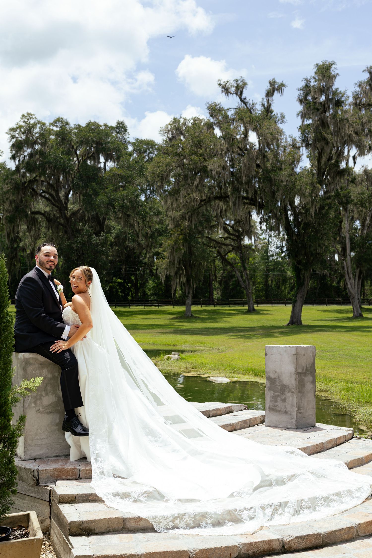Bride and groom pose on stone steps with long flowing veil, surrounded by moss-draped oak trees and green lawn.