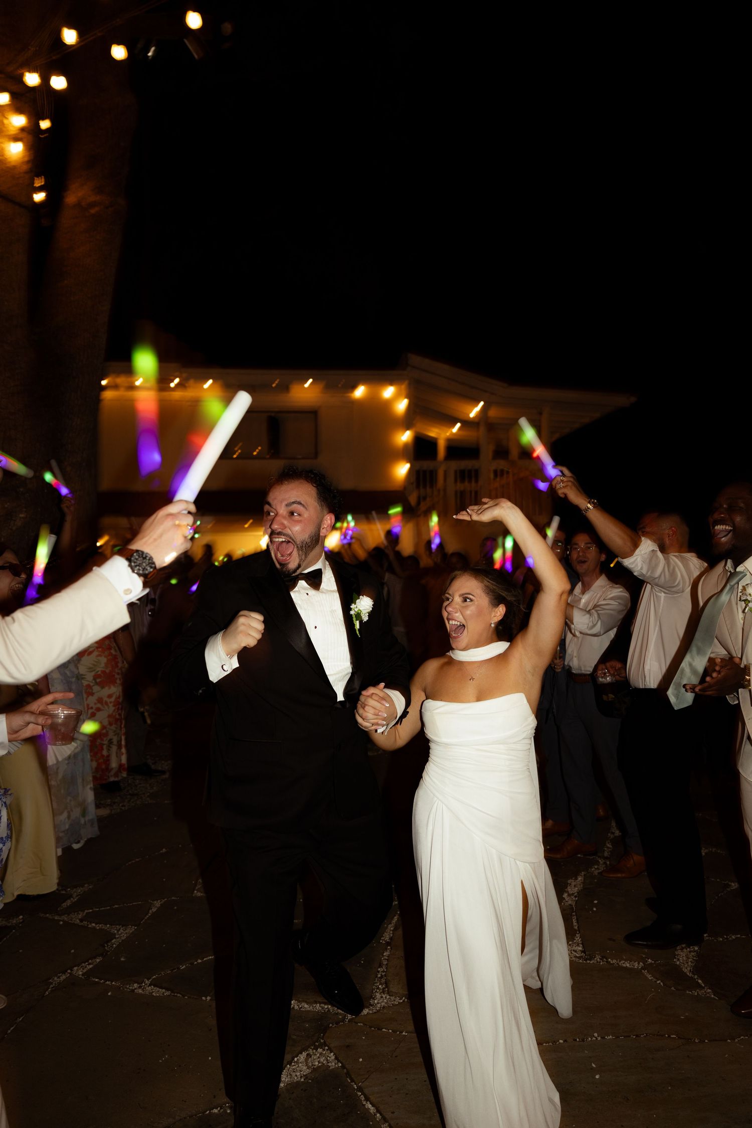 Bride and groom exit wedding reception at night surrounded by guests waving colorful glow sticks under string lights.