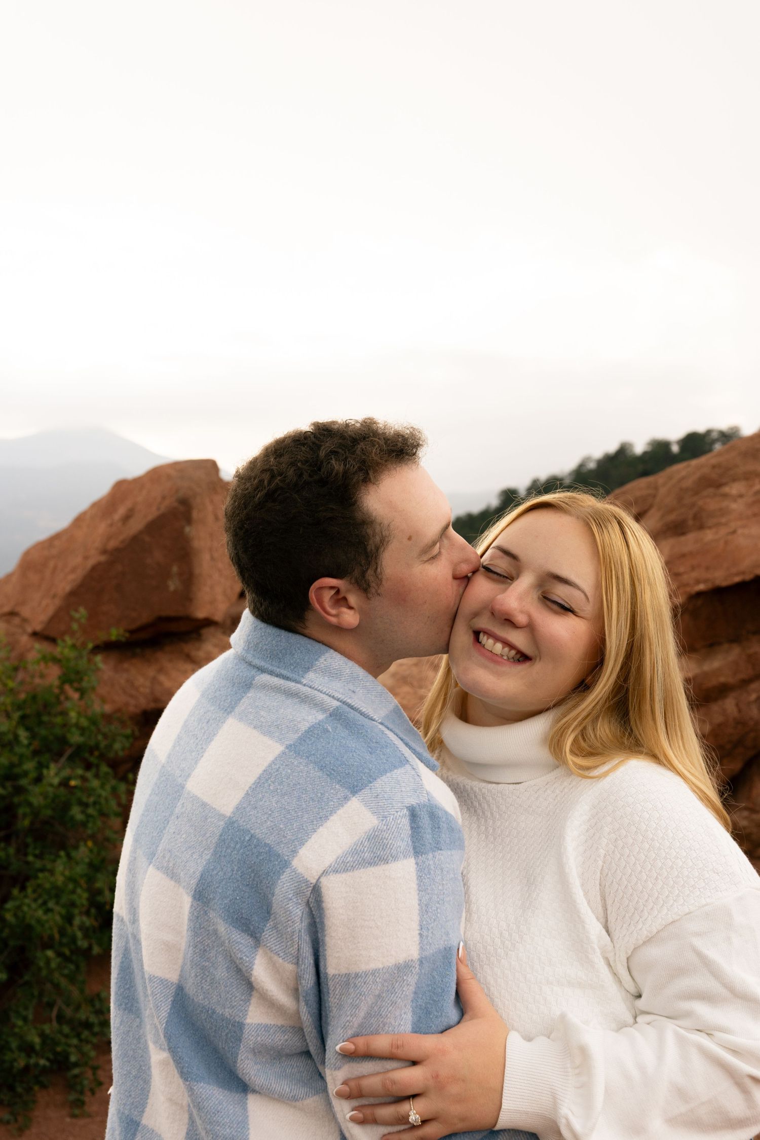 A couple shares a romantic kiss outdoors at Garden of the Gods, with red rock formations and mountains in the background.