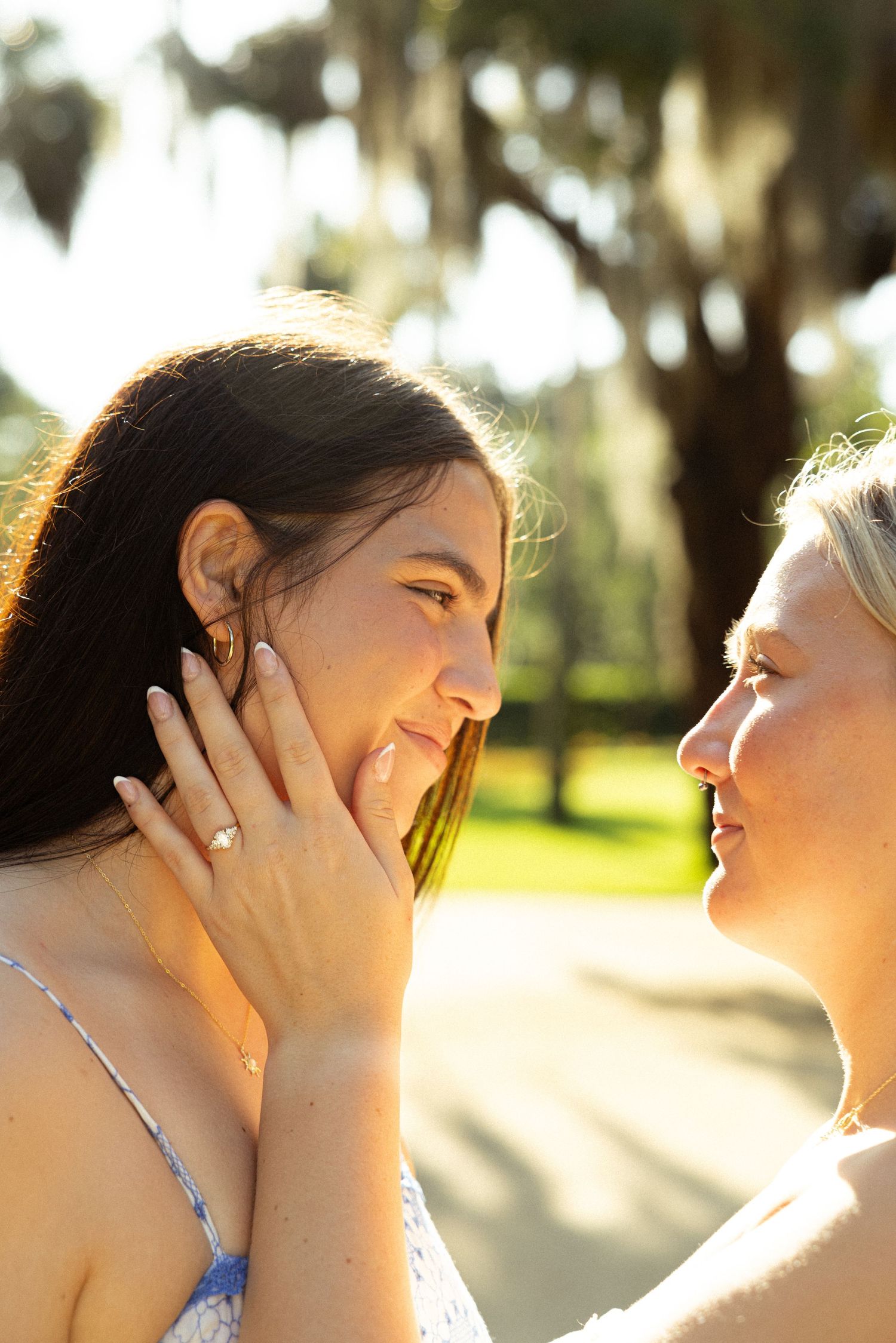 Two women share a tender moment outdoors, one gently cupping the other's face, smiling in a sunlit park.