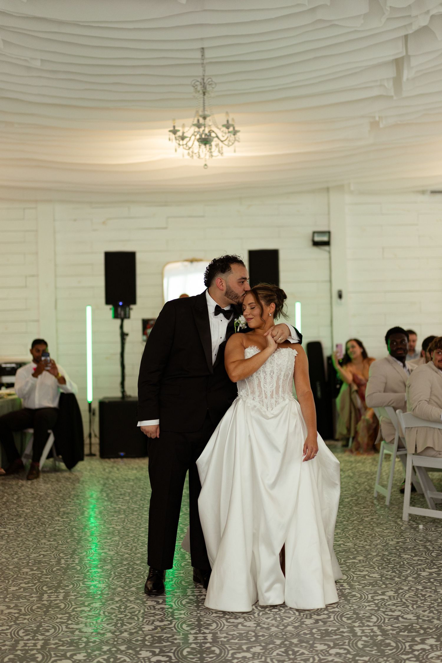 Bride and groom share first dance in elegant white ballroom with crystal chandeliers overhead.