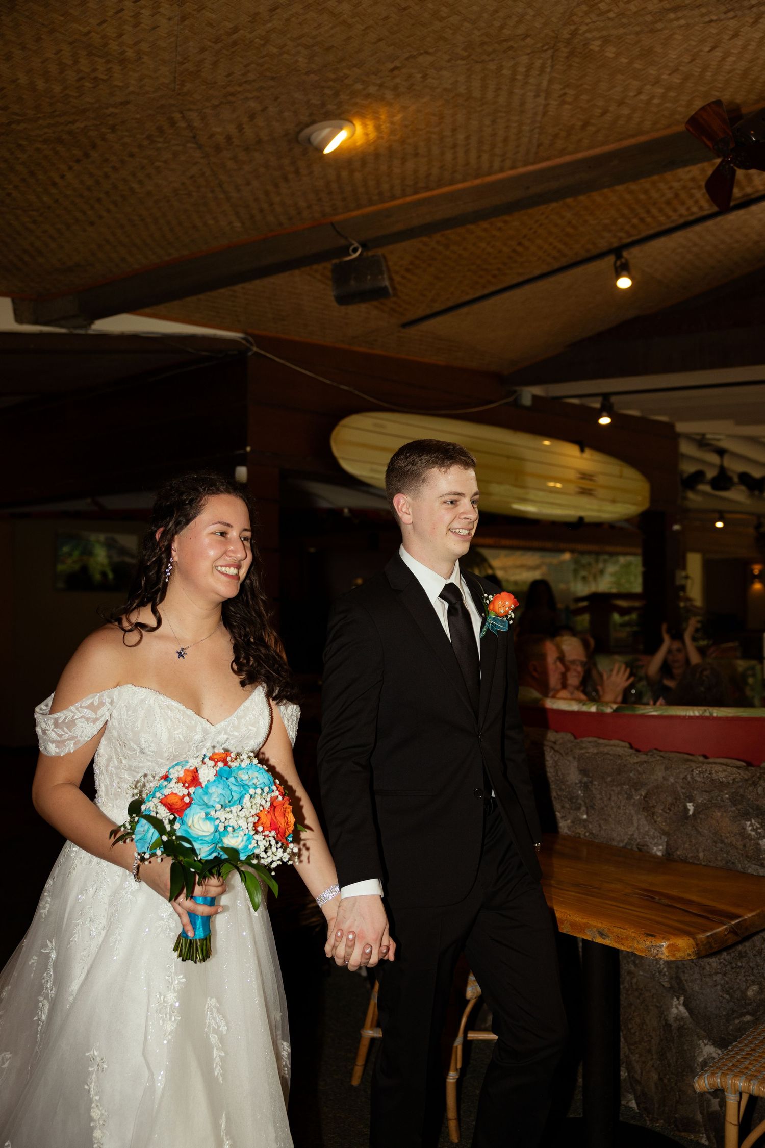 Smiling bride and groom walk hand-in-hand, she holds a blue and orange bouquet, he wears a black suit with boutonniere.