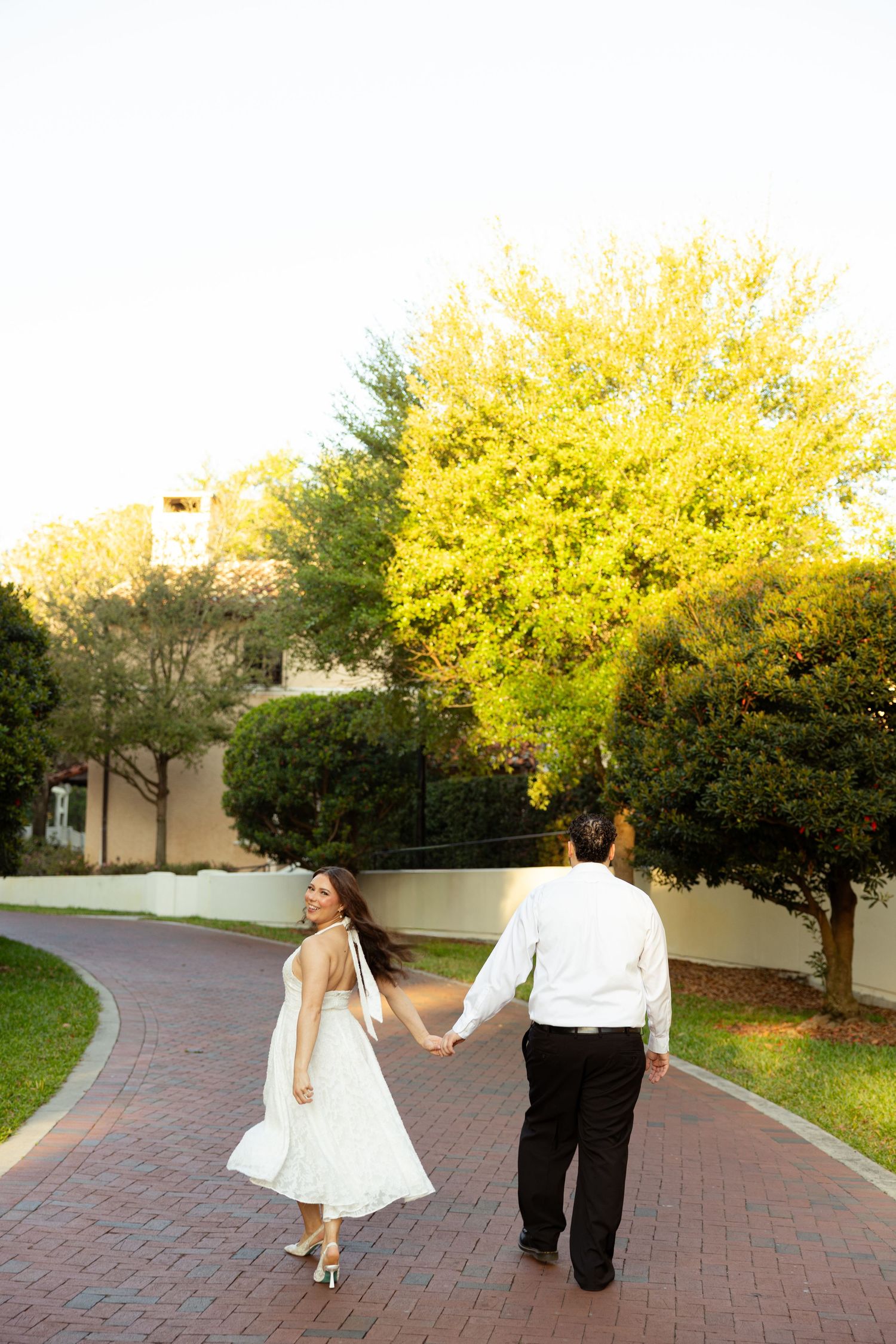 Couple holding hands walking on brick path in garden, woman in white dress, man in white shirt and black pants.