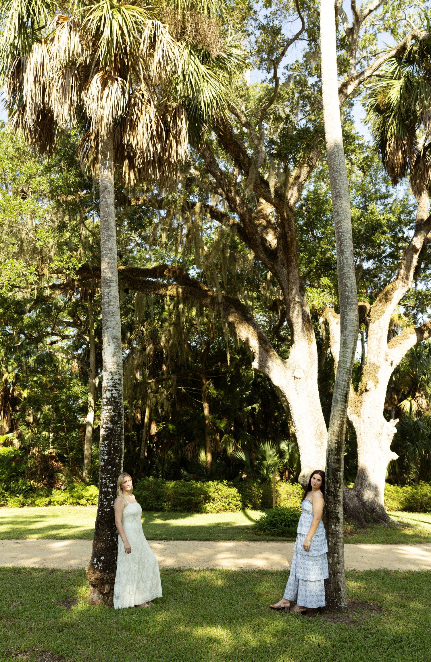 Two women in white dresses stand among tall palm trees in a lush green park on a sunny day.