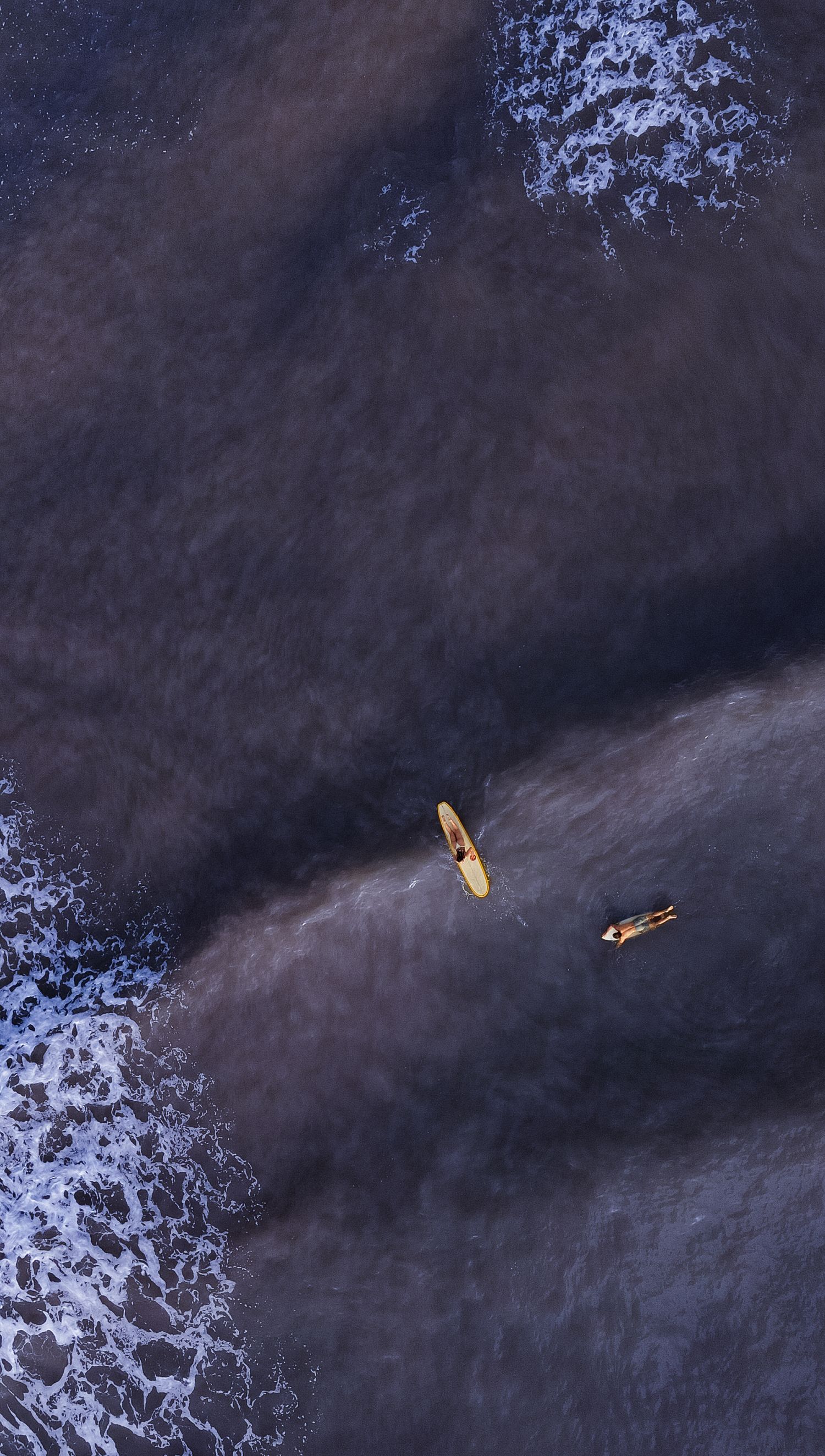 Aerial drone view of two surfers floating on dark ocean waves near a rocky shoreline.