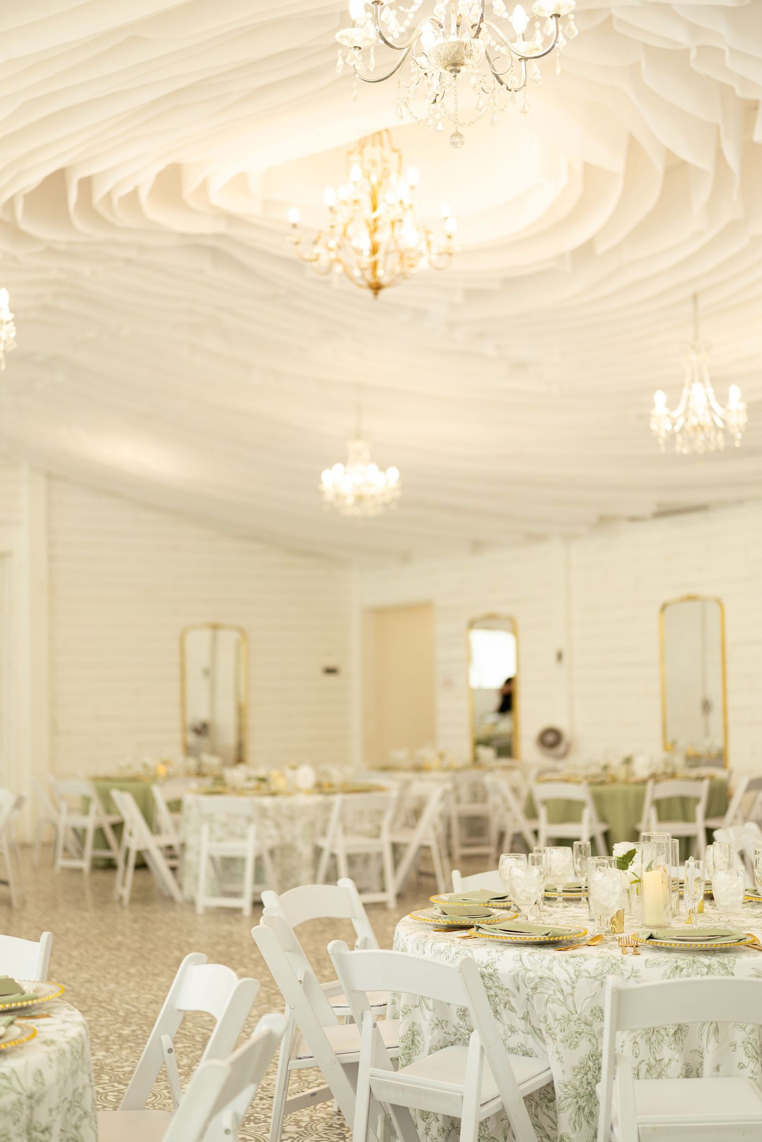Elegant white wedding reception hall with crystal chandeliers, floral tablecloths, and white chairs.