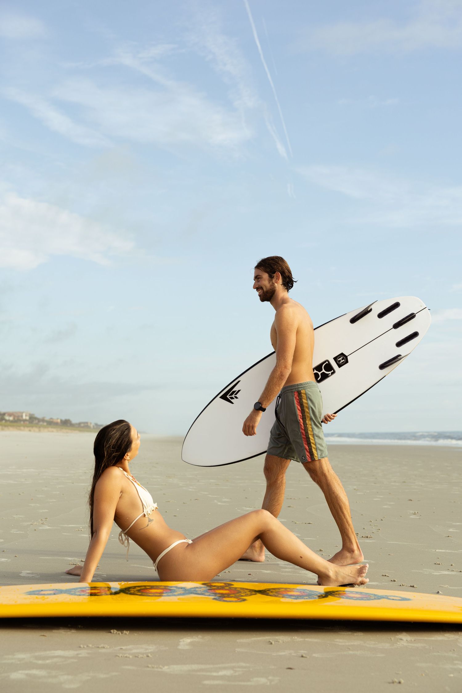 A man carrying a white surfboard walks past a woman in a bikini sitting on a yellow board on a sandy beach.