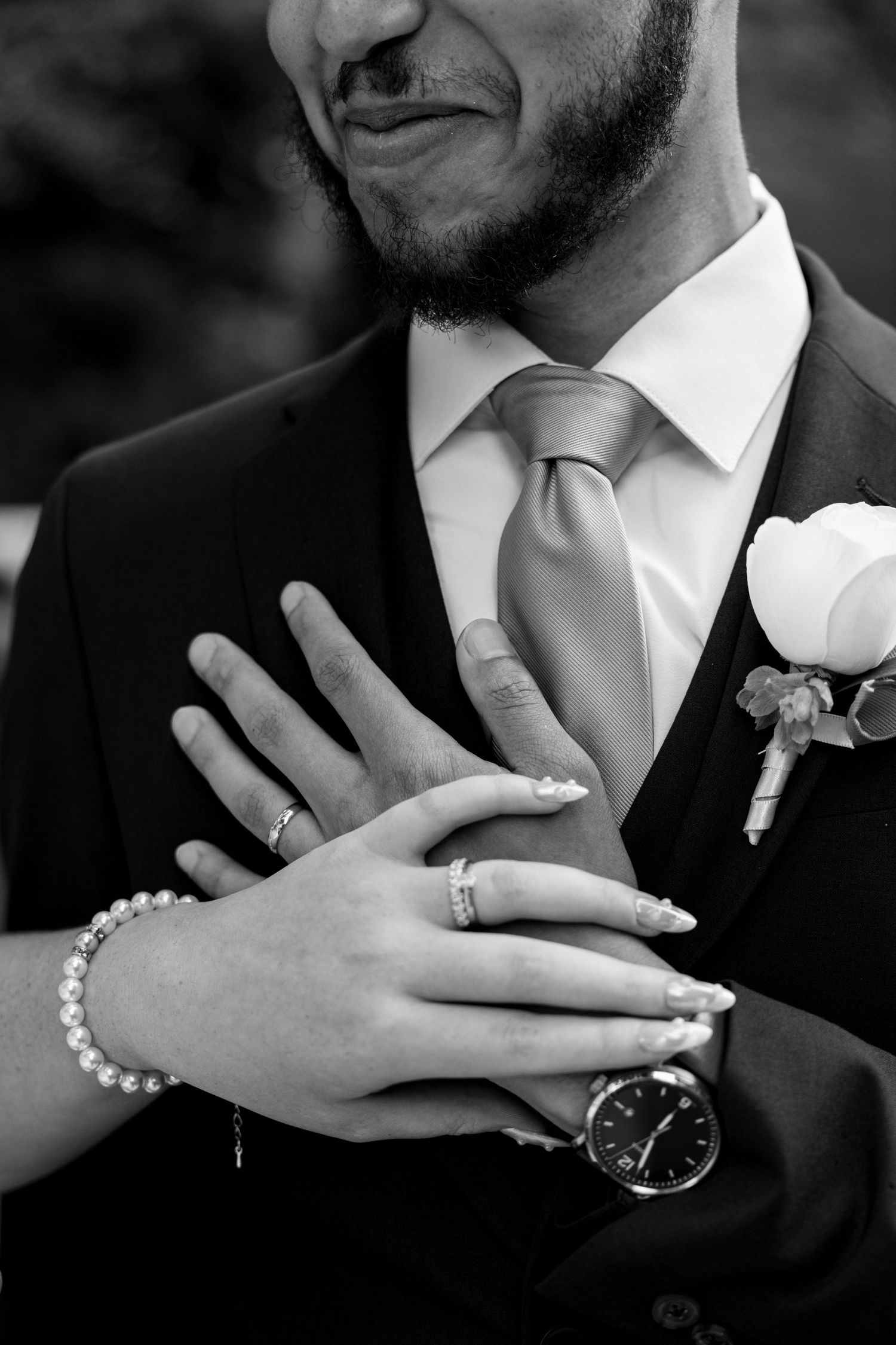 Black and white wedding photo of couple's hands with rings, pearl bracelet, and watch resting on groom's suited chest.