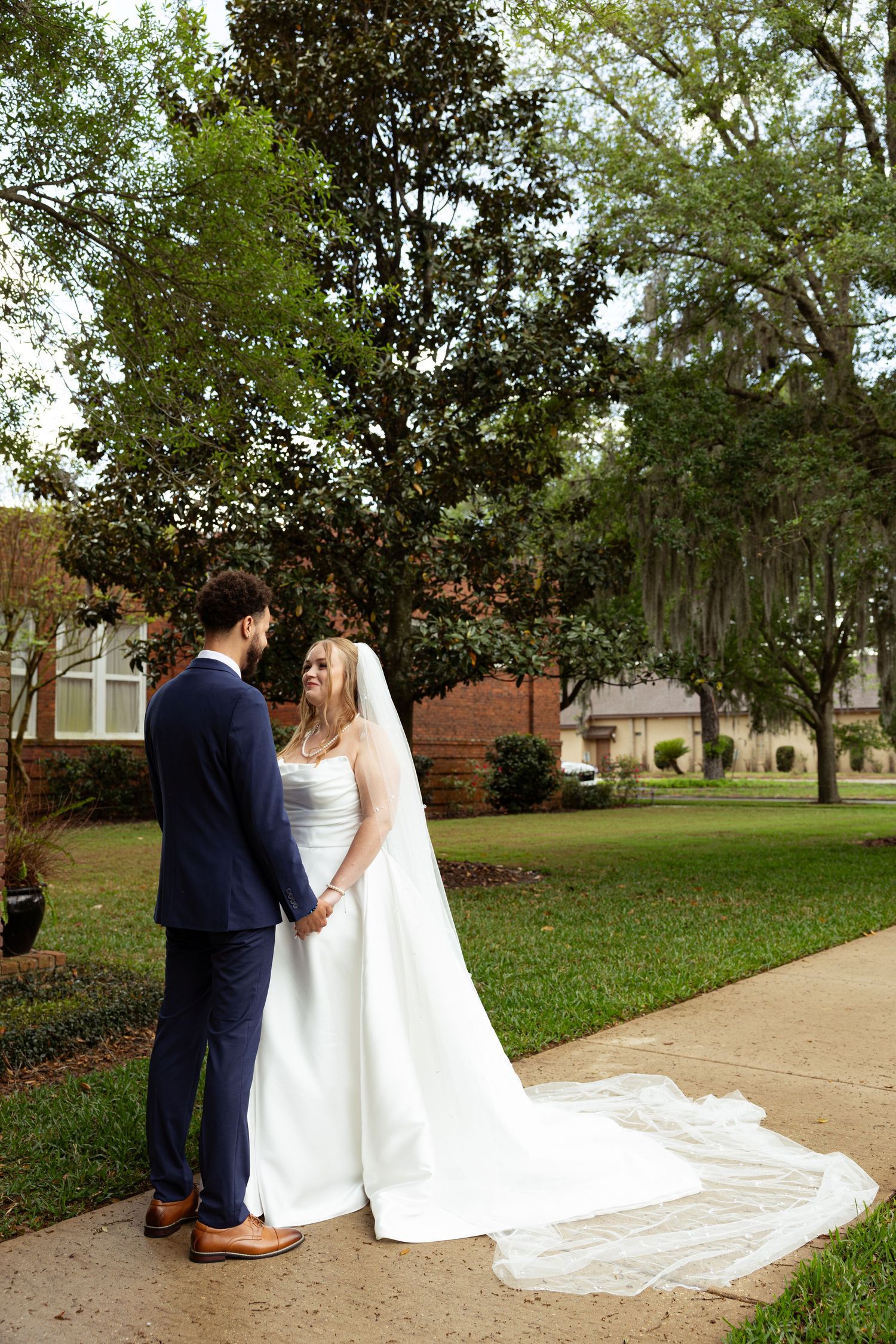 Bride and groom holding hands on a garden path, bride in white gown with long veil, groom in navy suit.