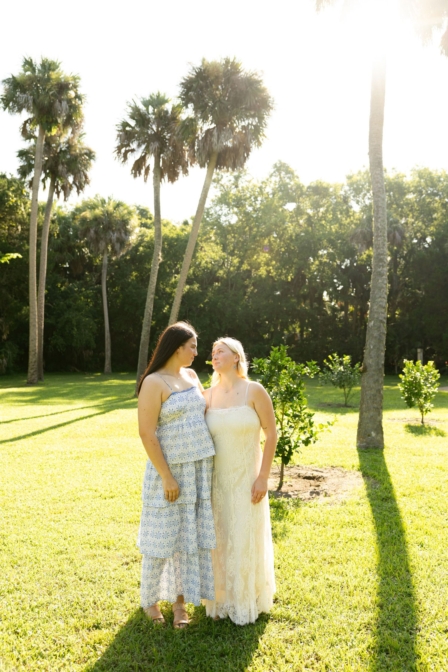 Two women in elegant dresses stand together on lush green grass surrounded by palm trees in warm sunlight.