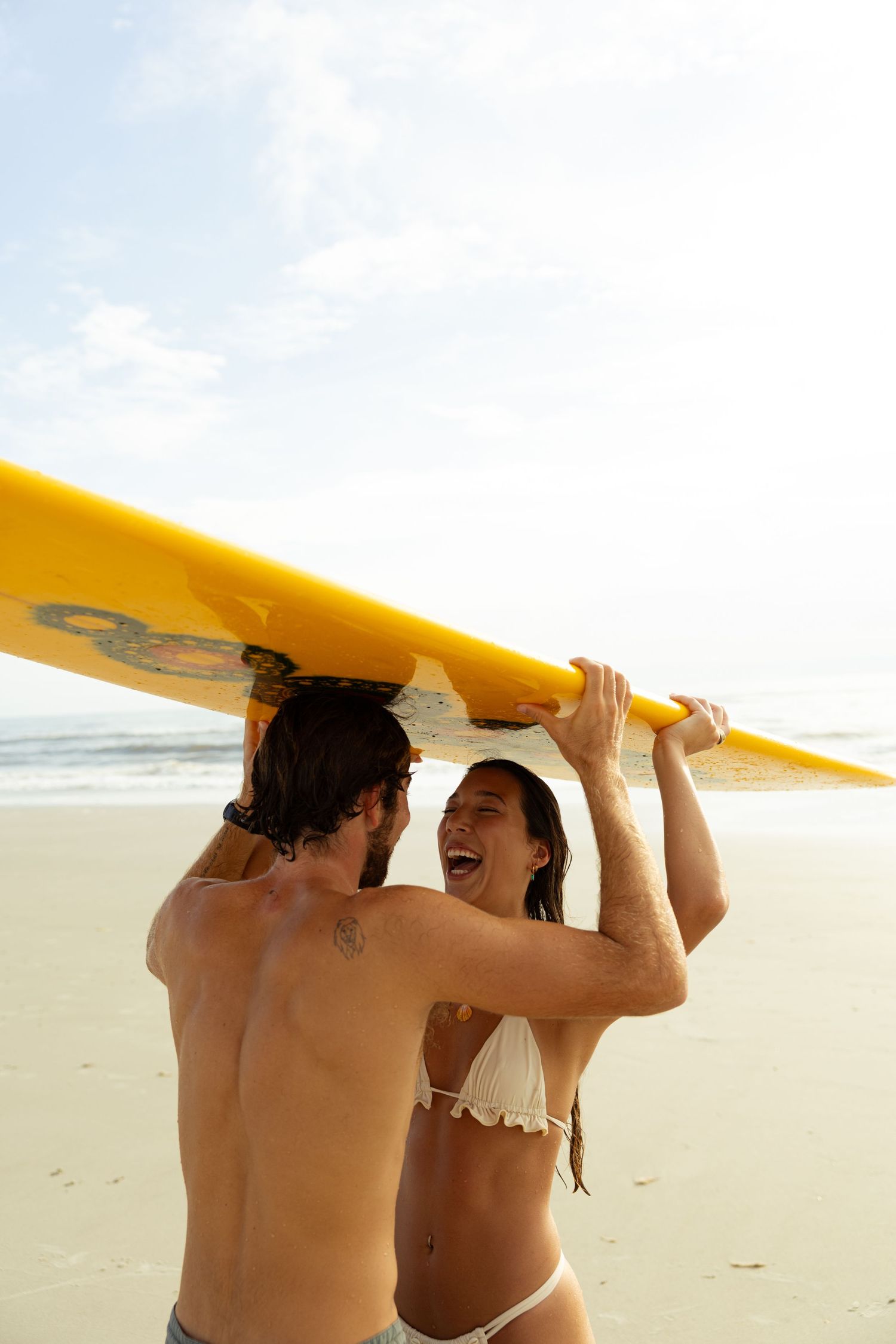 Couple laughing while carrying a yellow surfboard overhead on a sunny beach.