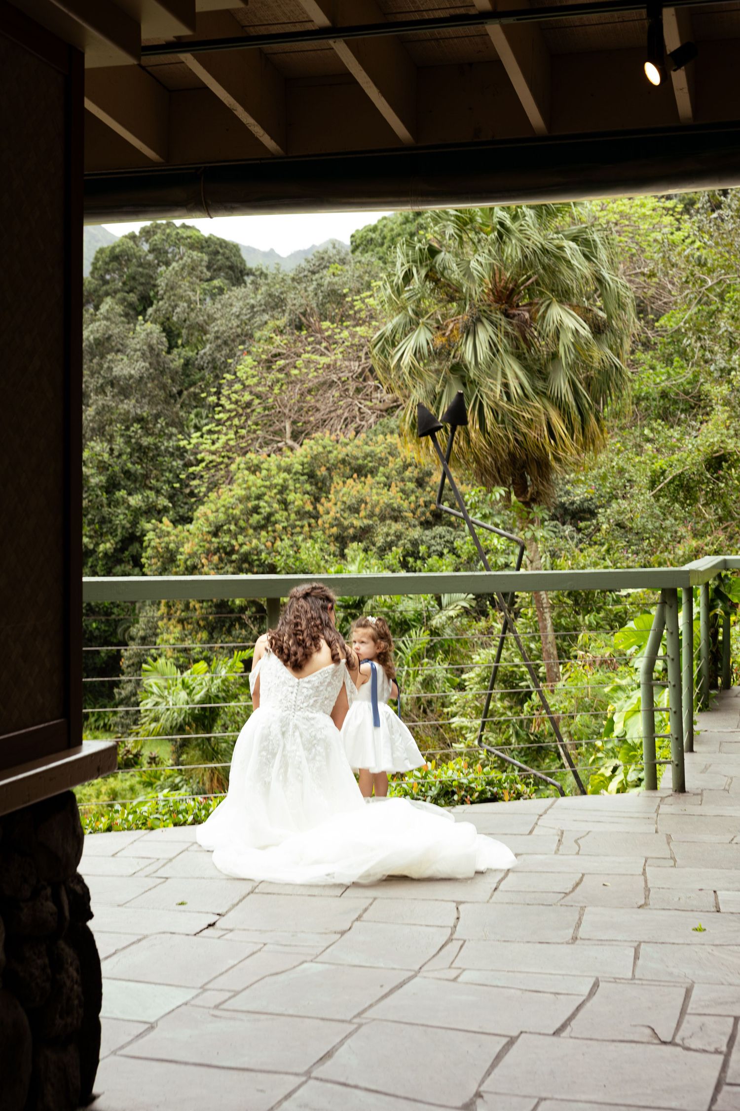 Bride in white gown kneels with flower girl on stone terrace overlooking lush tropical garden.