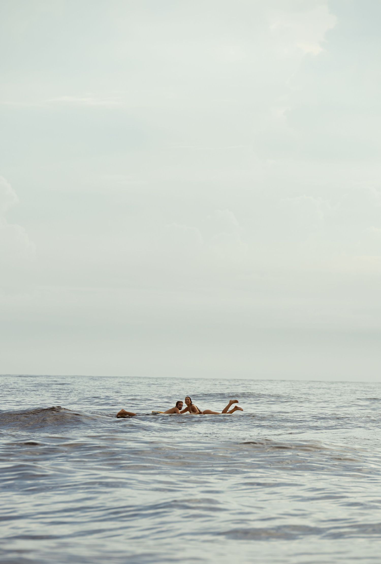 Two swimmers floating in calm ocean waters under a soft, hazy sky on a peaceful day.