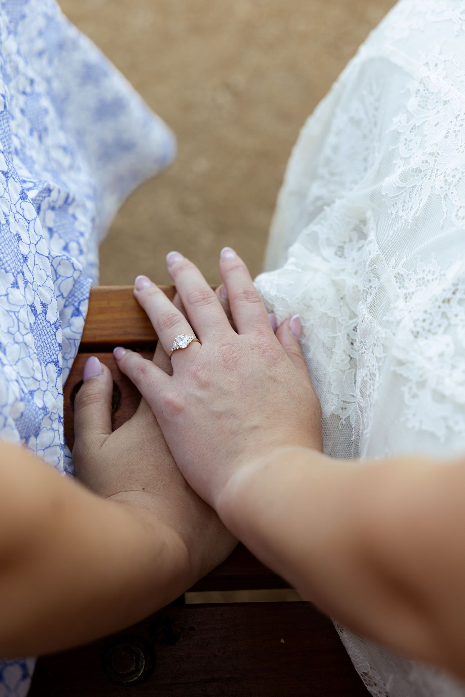 Bride and bridesmaid holding hands, showing off diamond engagement ring, wearing white lace and blue floral dresses.