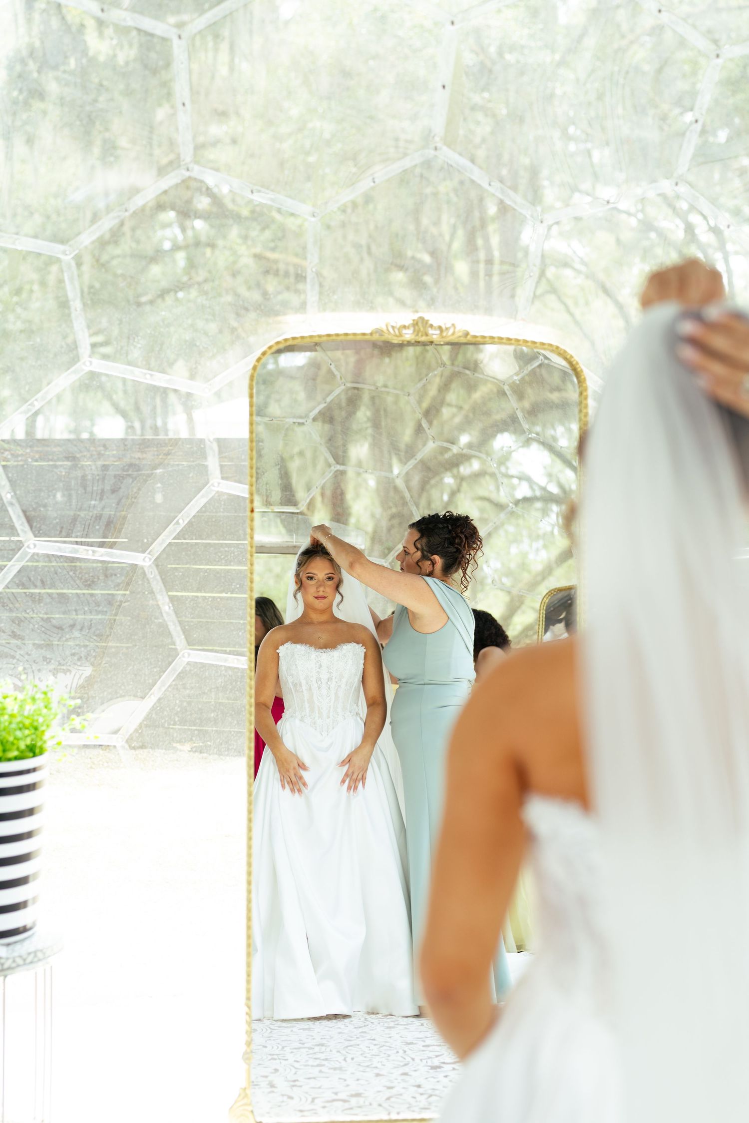 Bridesmaid adjusts brides veil reflected in ornate gold mirror inside bright wedding venue.