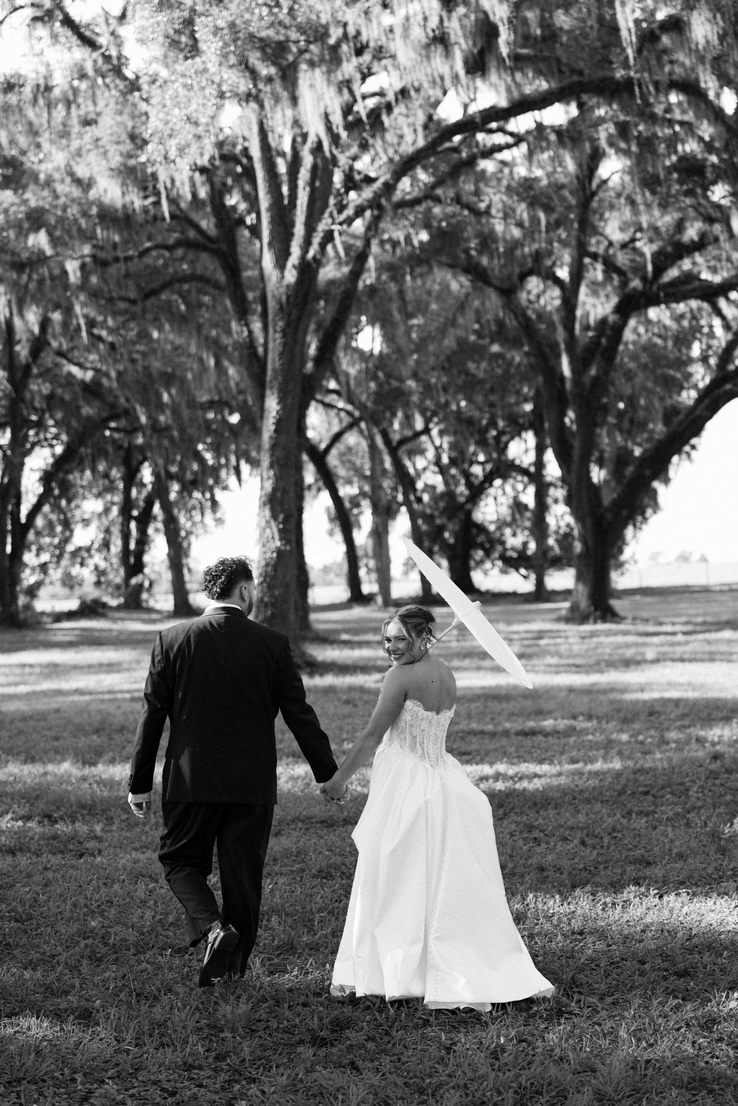 Black and white photo of a bride and groom walking hand in hand through a tree-lined lawn on their wedding day.