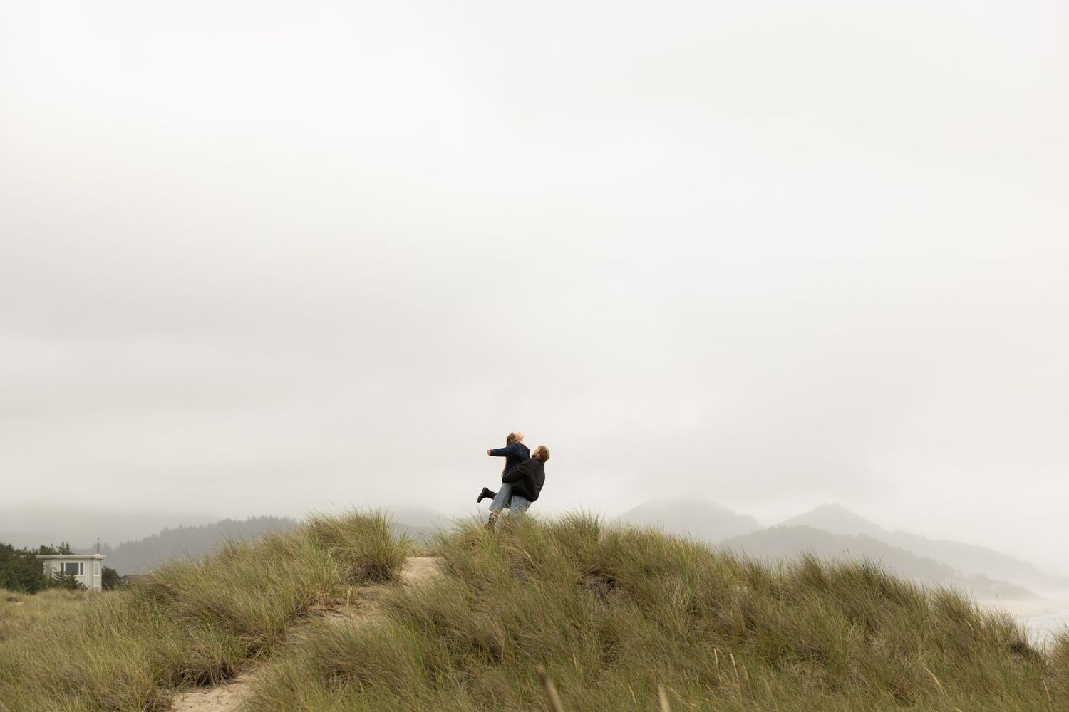 Person jumping on a grassy sand dune under an overcast sky with misty mountains in the background.