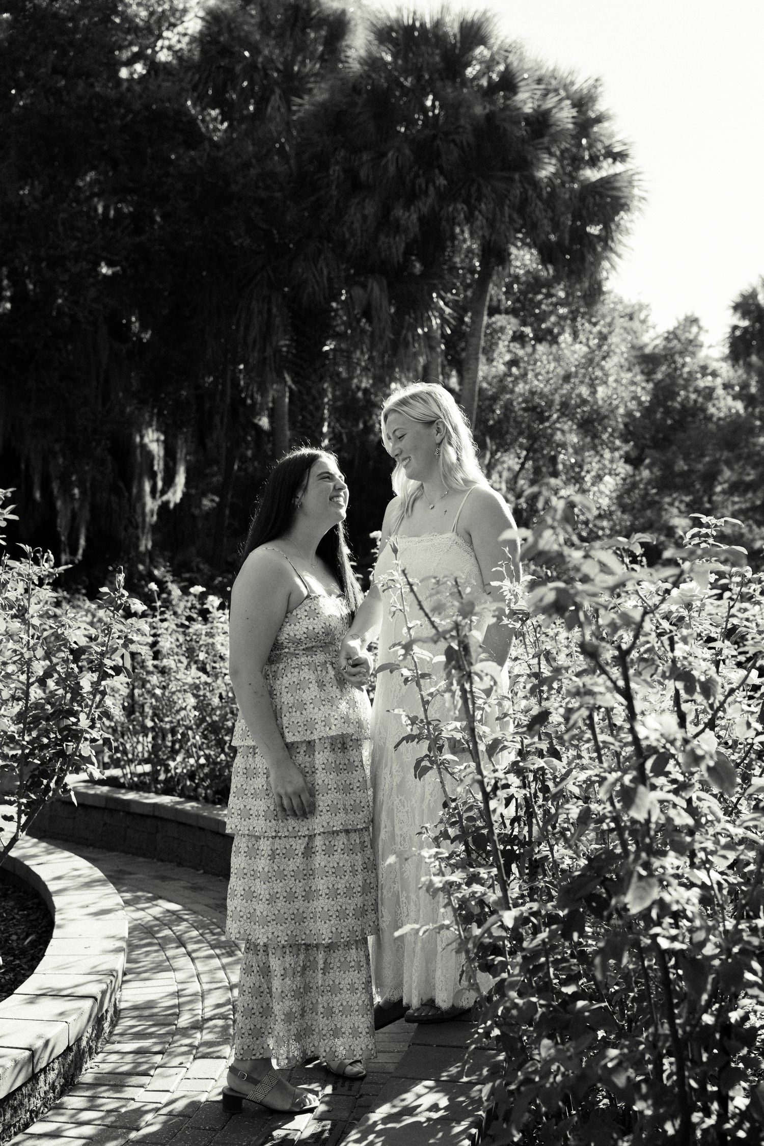 Two women in dresses stand together in a lush garden path, sharing a moment in black and white photography.