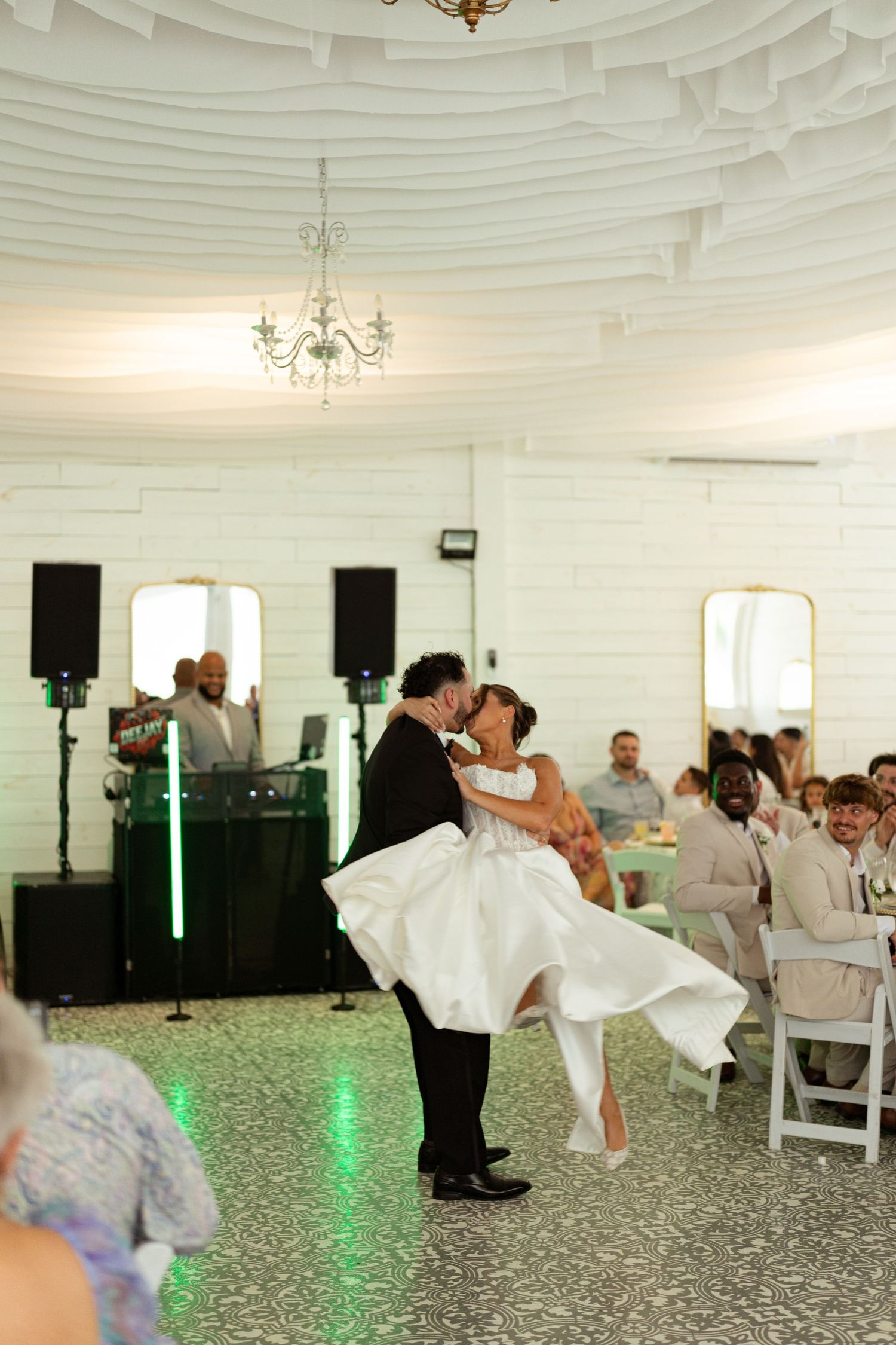 Groom lifts bride during first dance in elegant white domed reception hall with guests watching.