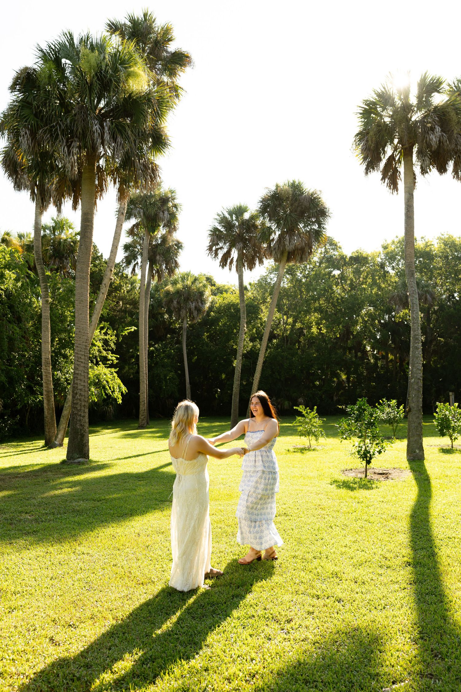 Two women in summer dresses hold hands and dance on a sunny green lawn surrounded by tall palm trees.