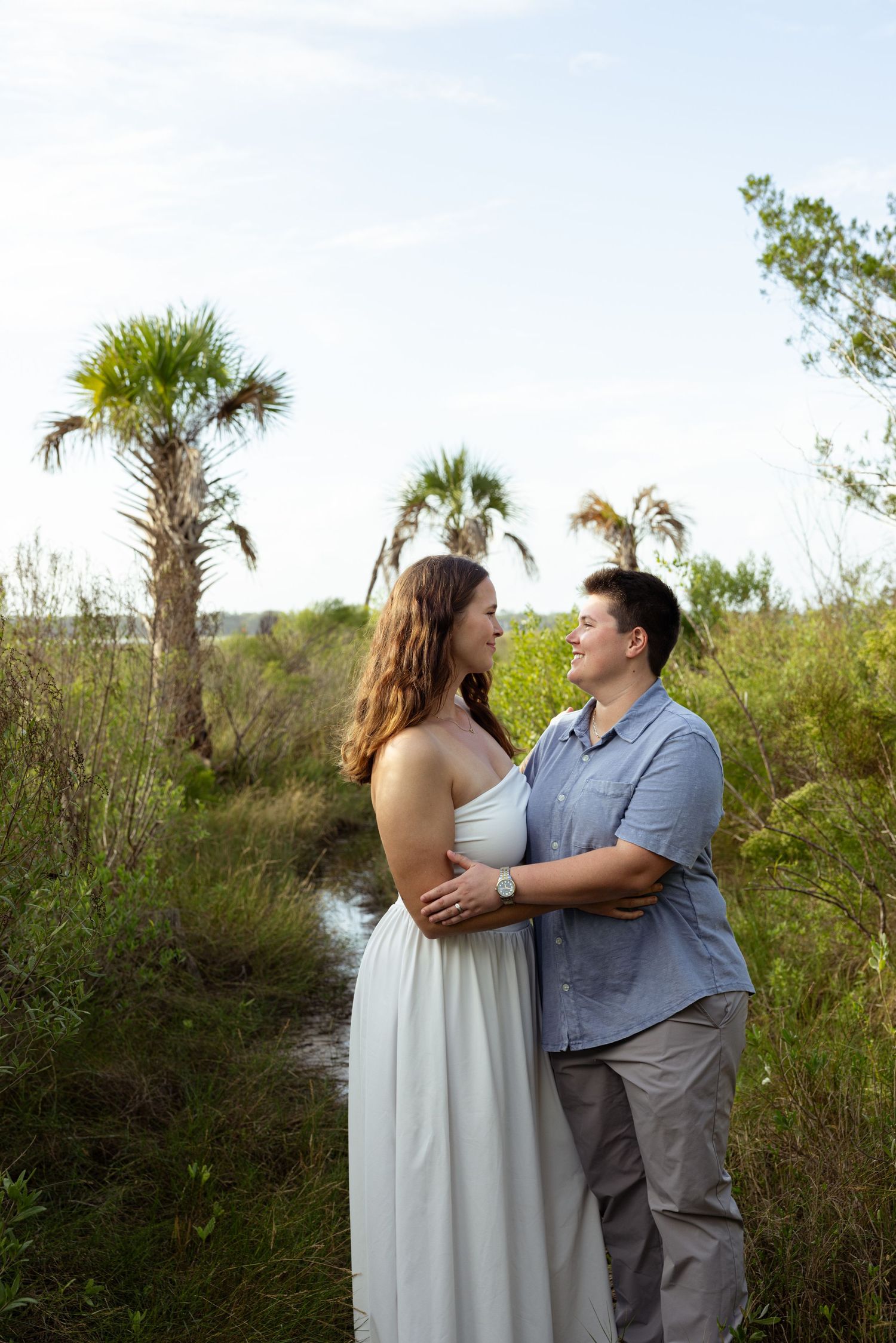 Couple embracing outdoors near palm trees and lush greenery, woman in white dress, man in blue shirt, smiling at each other.
