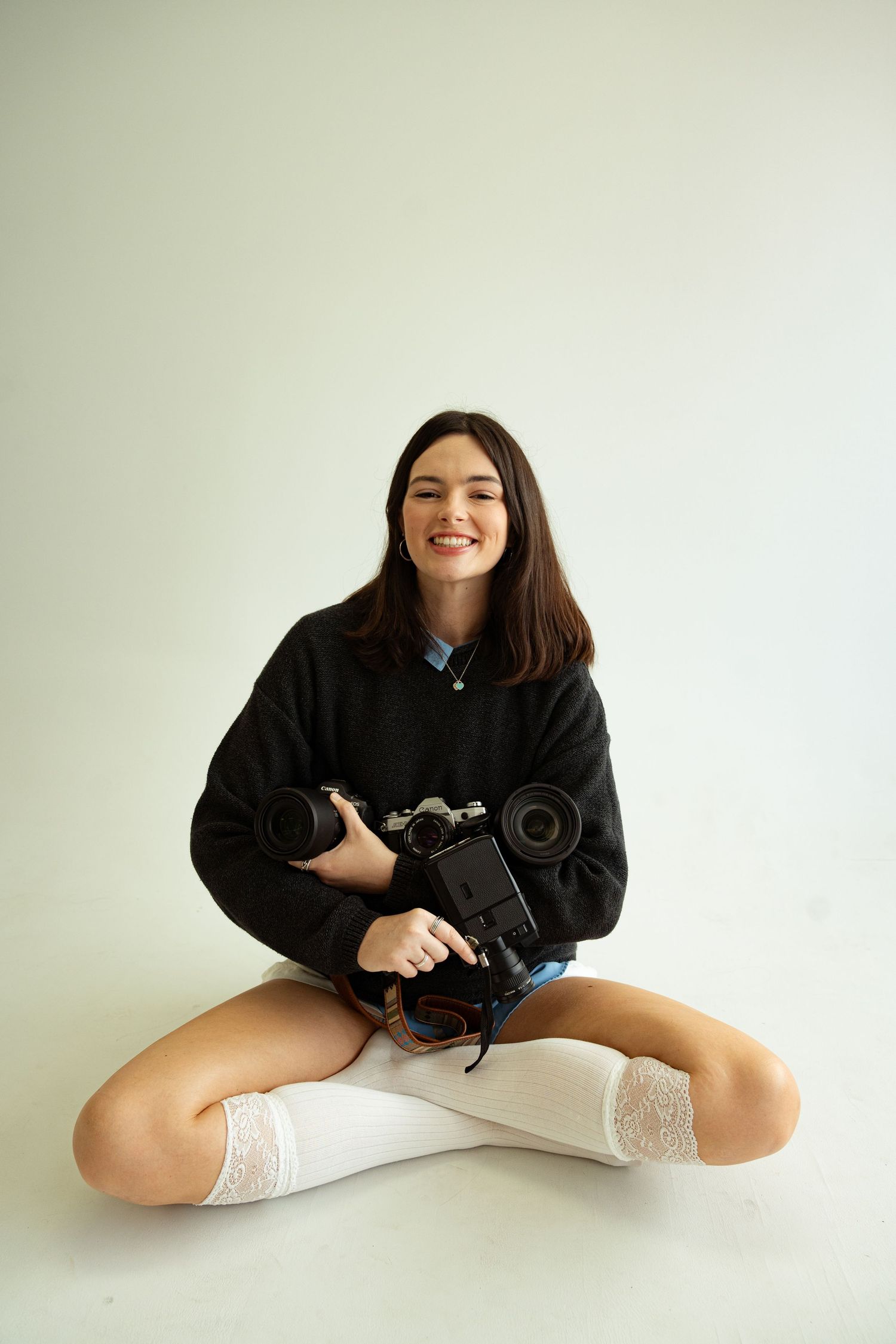 Smiling young woman sitting cross-legged on white floor holding camera and lens, wearing black sweater and white knee socks.