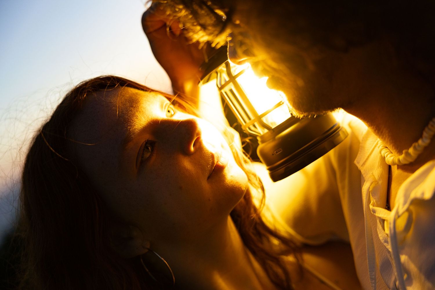 A woman gazes up at a glowing lantern held above her face, bathed in warm golden light at dusk.