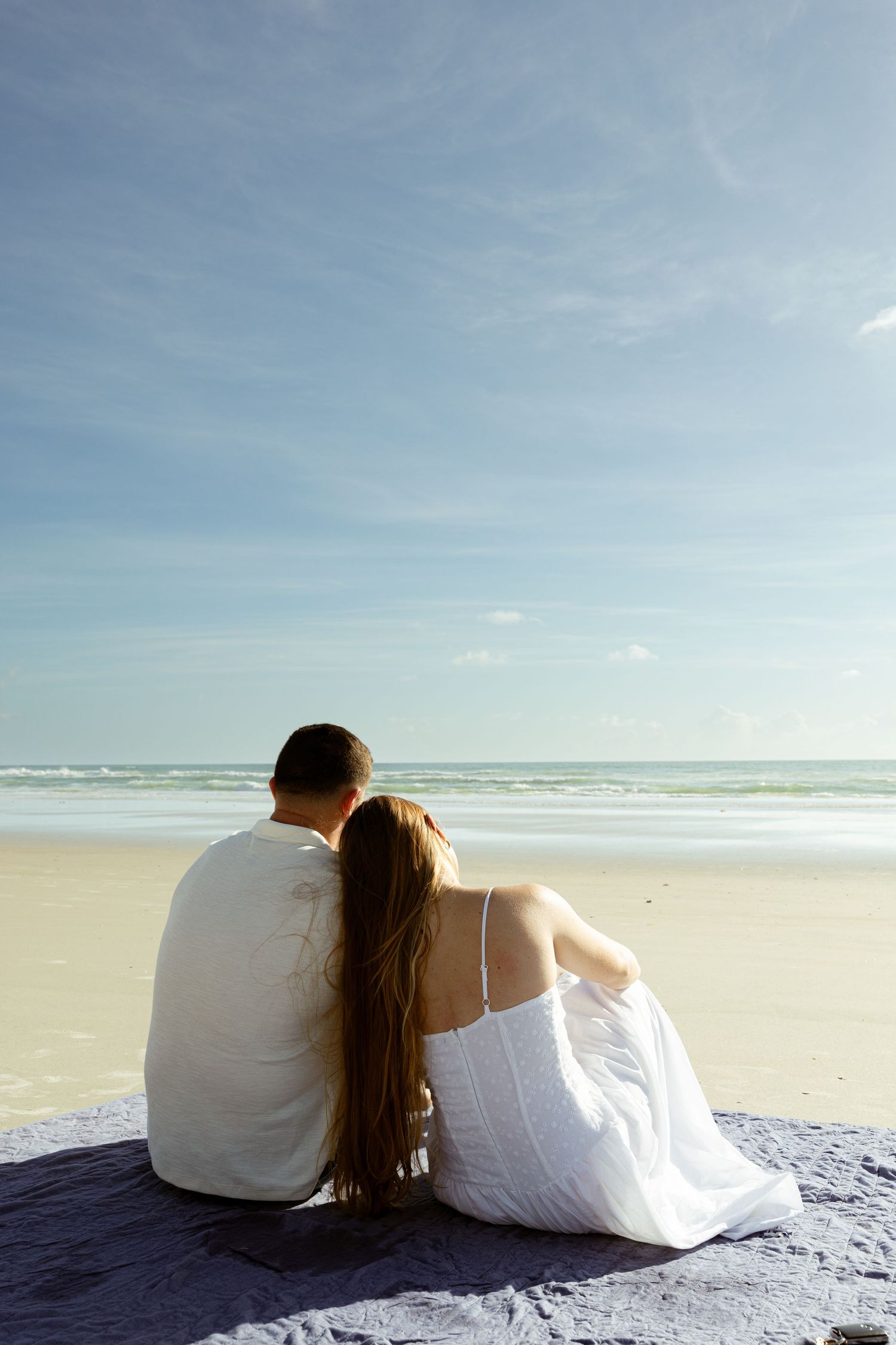Couple sitting together on a beach blanket, gazing at the ocean waves under a blue sky.