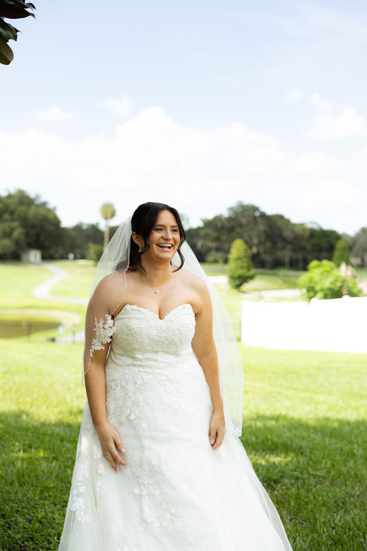 Smiling bride in strapless lace mermaid wedding gown and veil poses outdoors on green lawn.