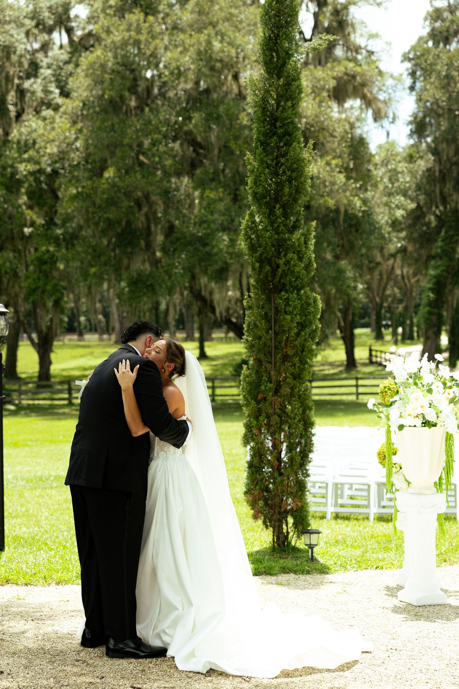 Bride and groom share first kiss at outdoor wedding ceremony beside tall cypress tree and floral arrangements.