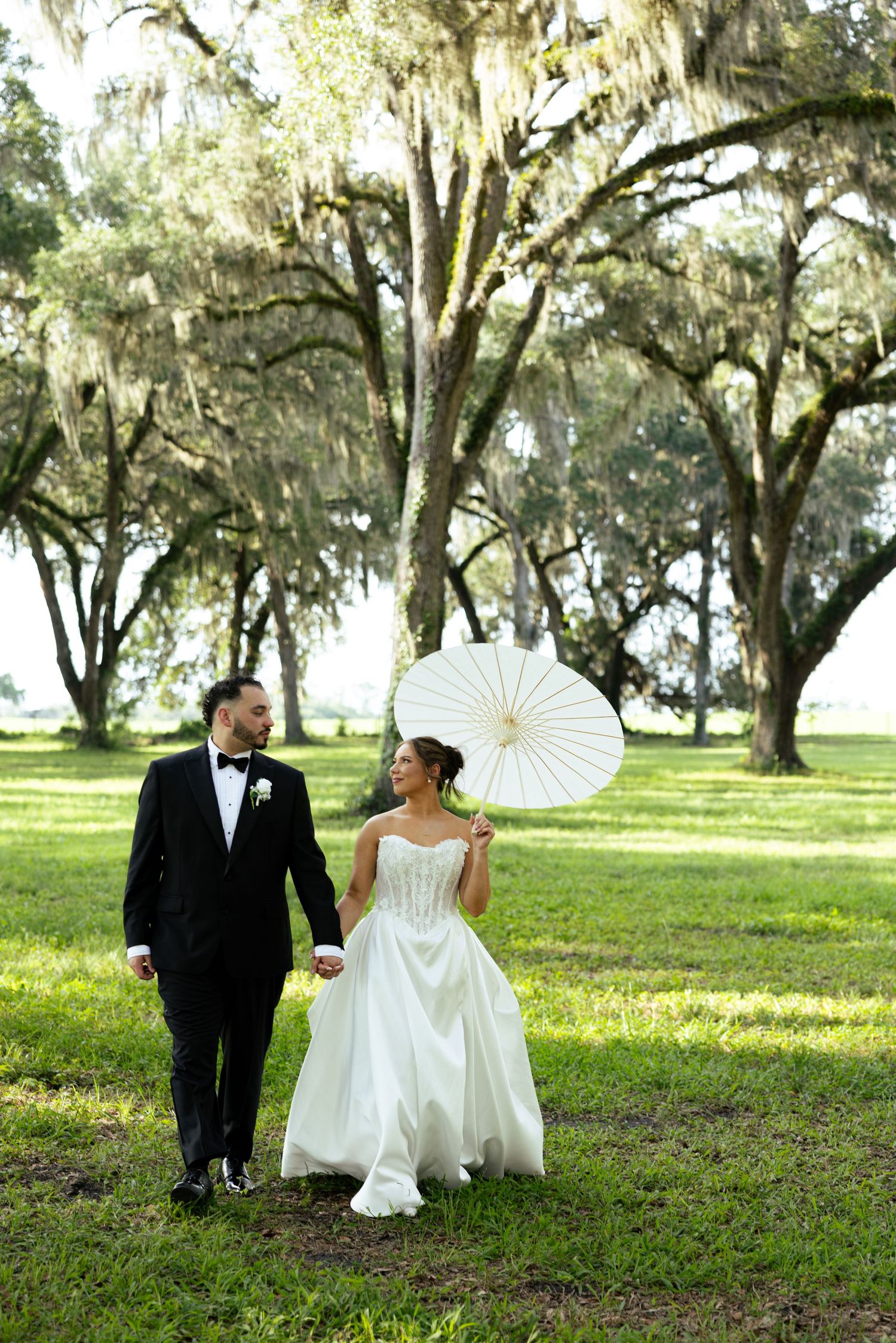 Bride in strapless gown holds white parasol while walking hand-in-hand with groom under Spanish moss-draped oak trees.