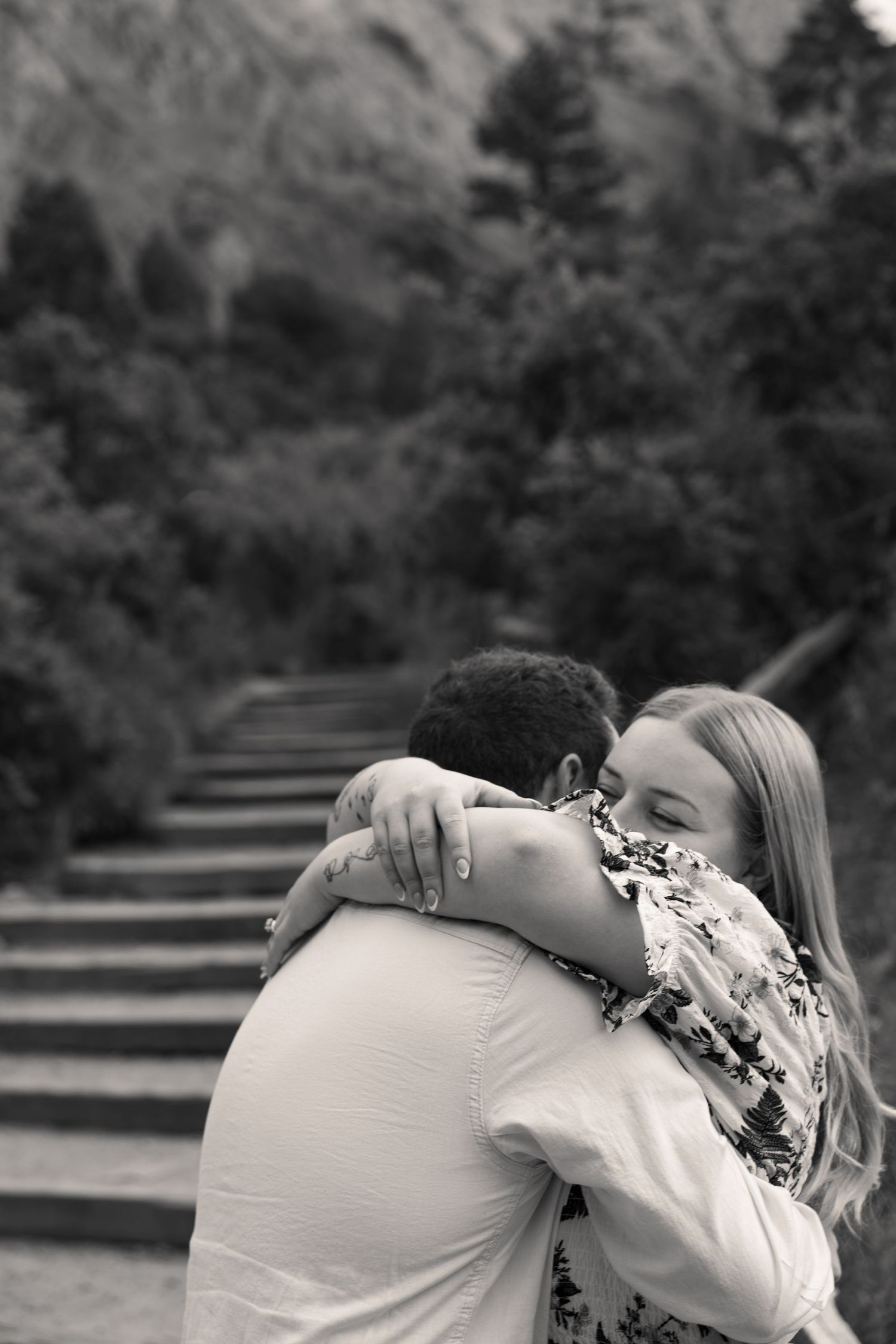 Black and white photo of a couple embracing and smiling on outdoor stairs surrounded by lush green trees.