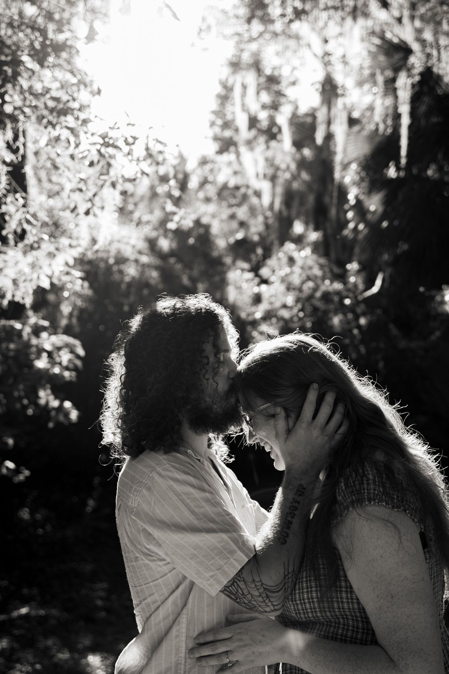 Romantic black and white photo of a couple almost kissing in a sun-dappled forest setting.