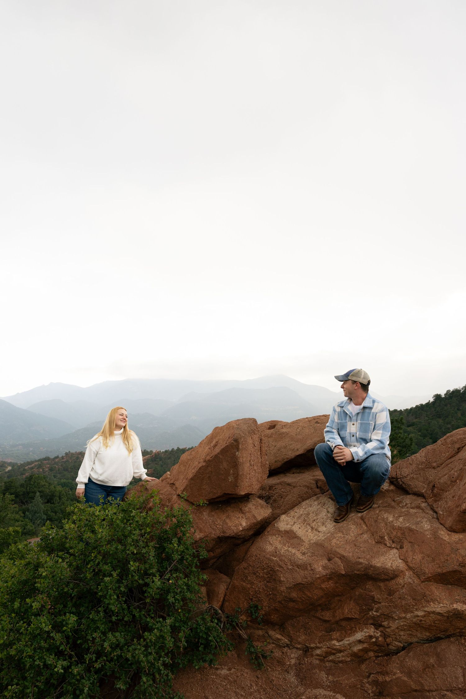 Couple sitting on red rocks at Garden of the Gods with panoramic mountain views on an overcast day.