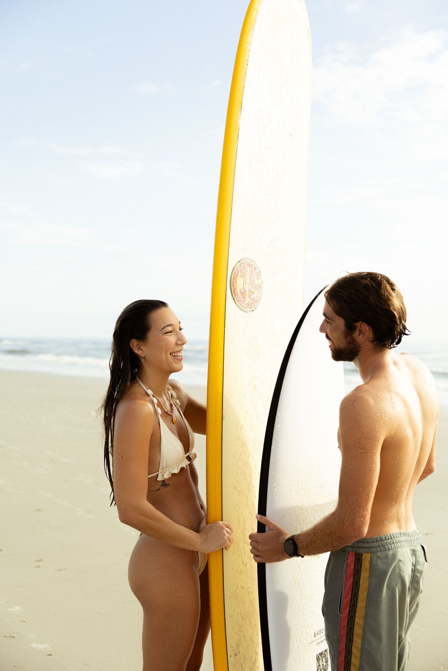 A smiling woman in a bikini and a shirtless man hold a yellow surfboard together on a sunny beach.