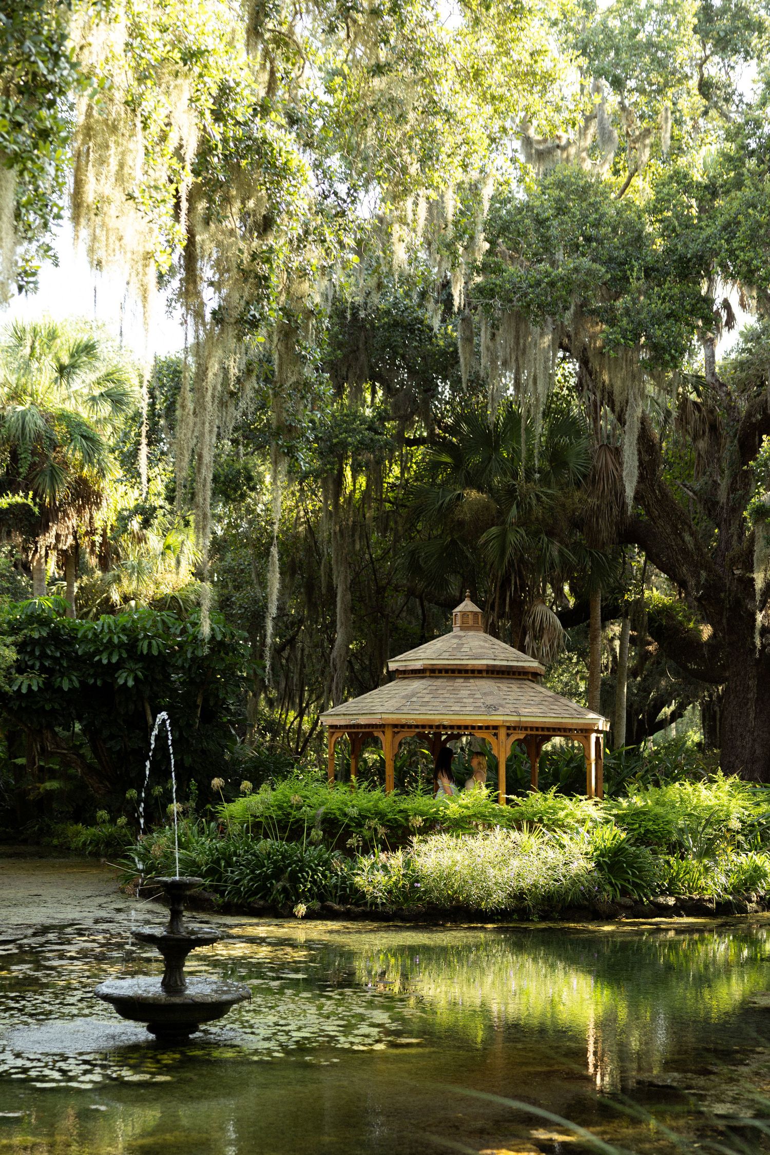 A serene garden gazebo reflects in a lily pond with a fountain, surrounded by lush greenery and Spanish moss-draped trees.