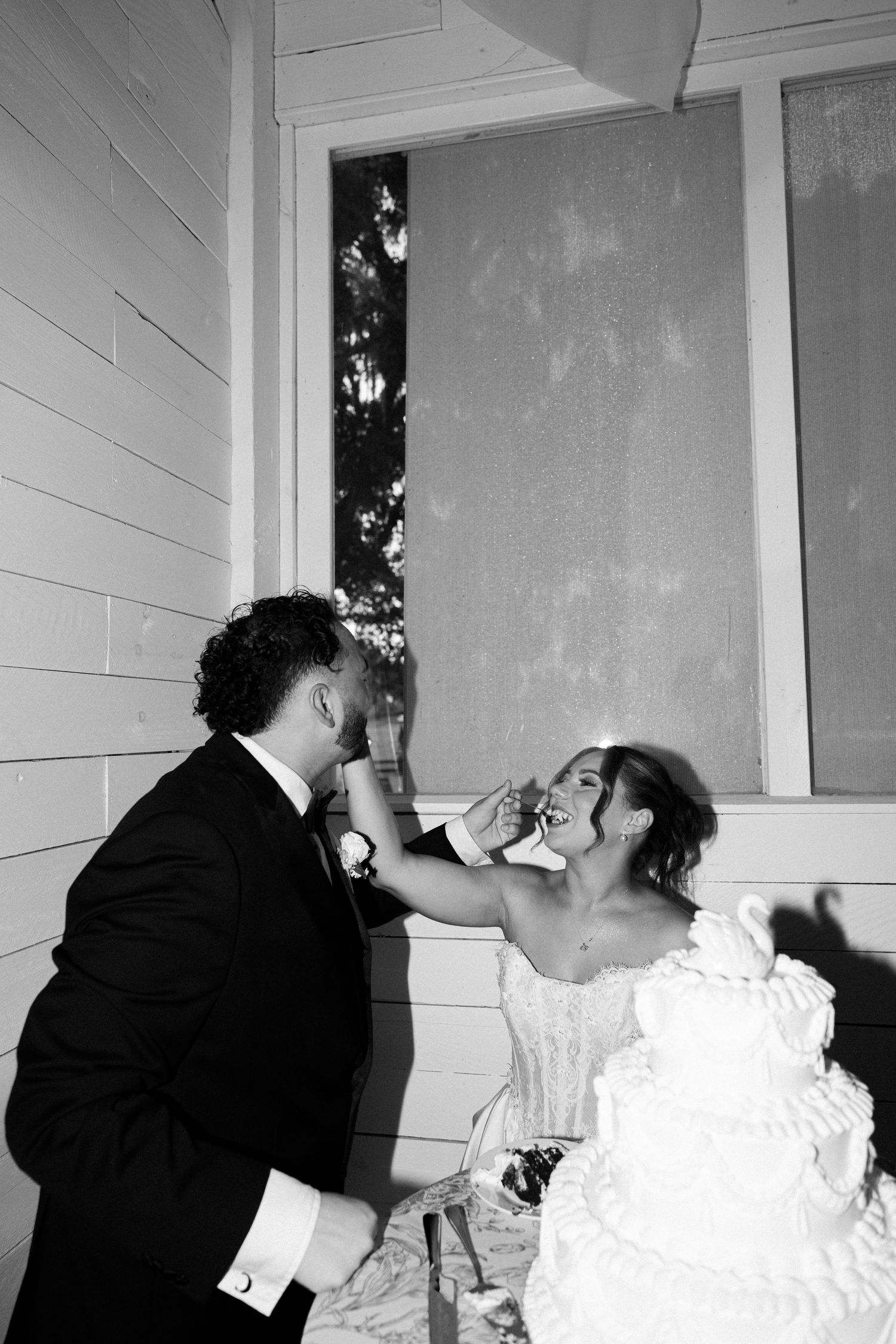 Bride playfully feeds groom wedding cake in a joyful black and white outdoor reception photo.