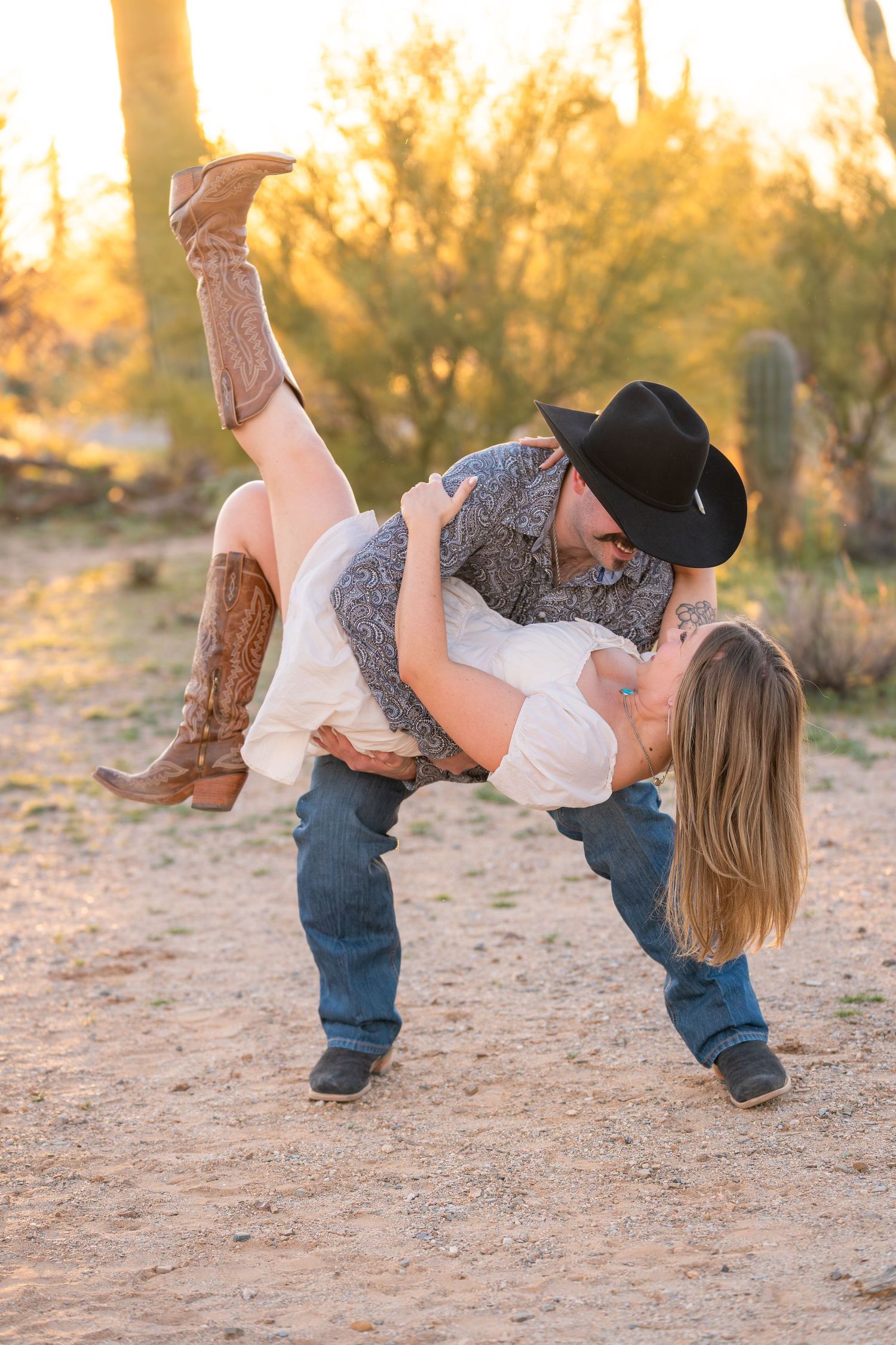 Playful Western engagement photo in Arizona desert with cowboy dipping cowgirl at golden hour