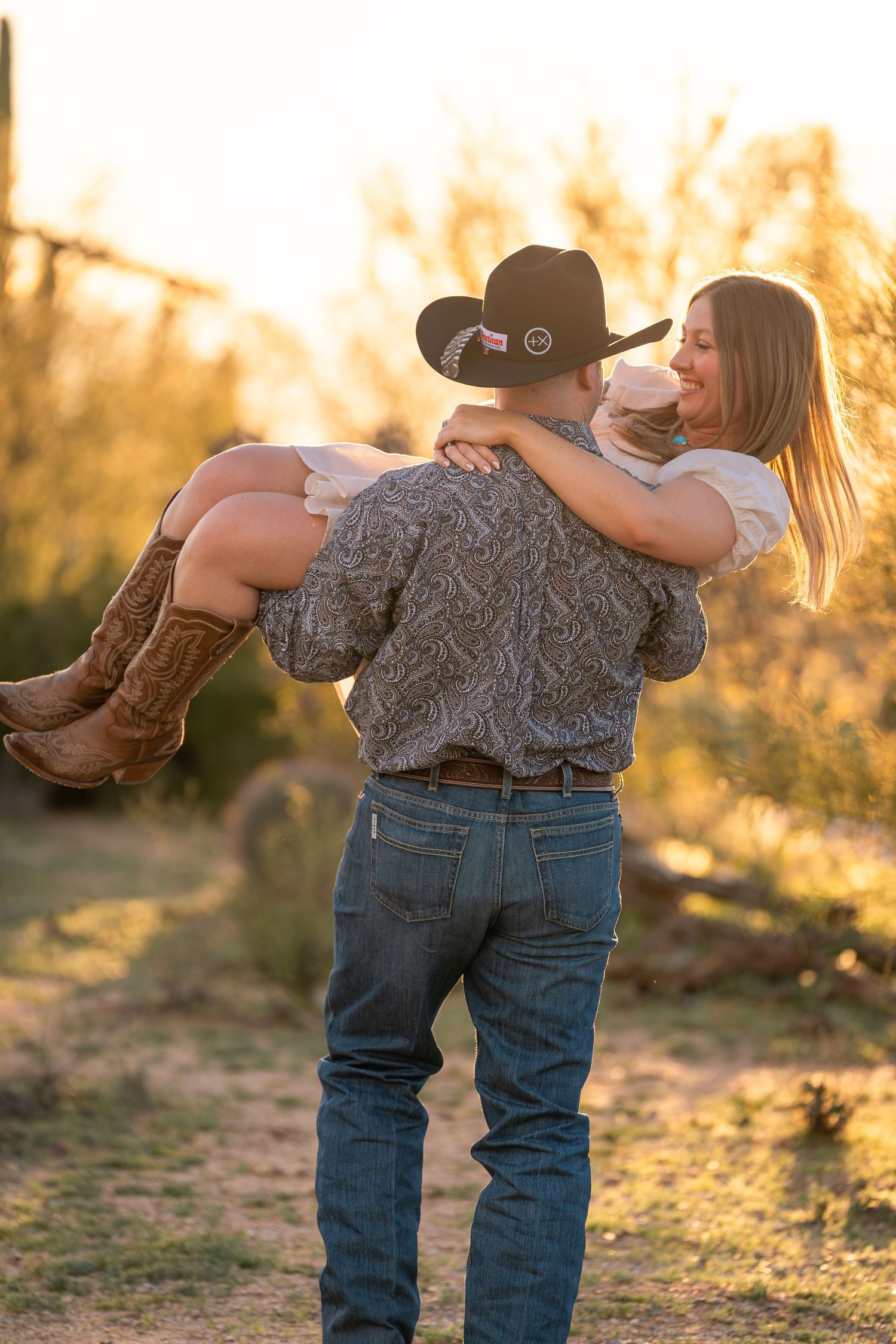 Western couple at sunset in Arizona desert, cowboy lifting cowgirl during golden hour engagement session