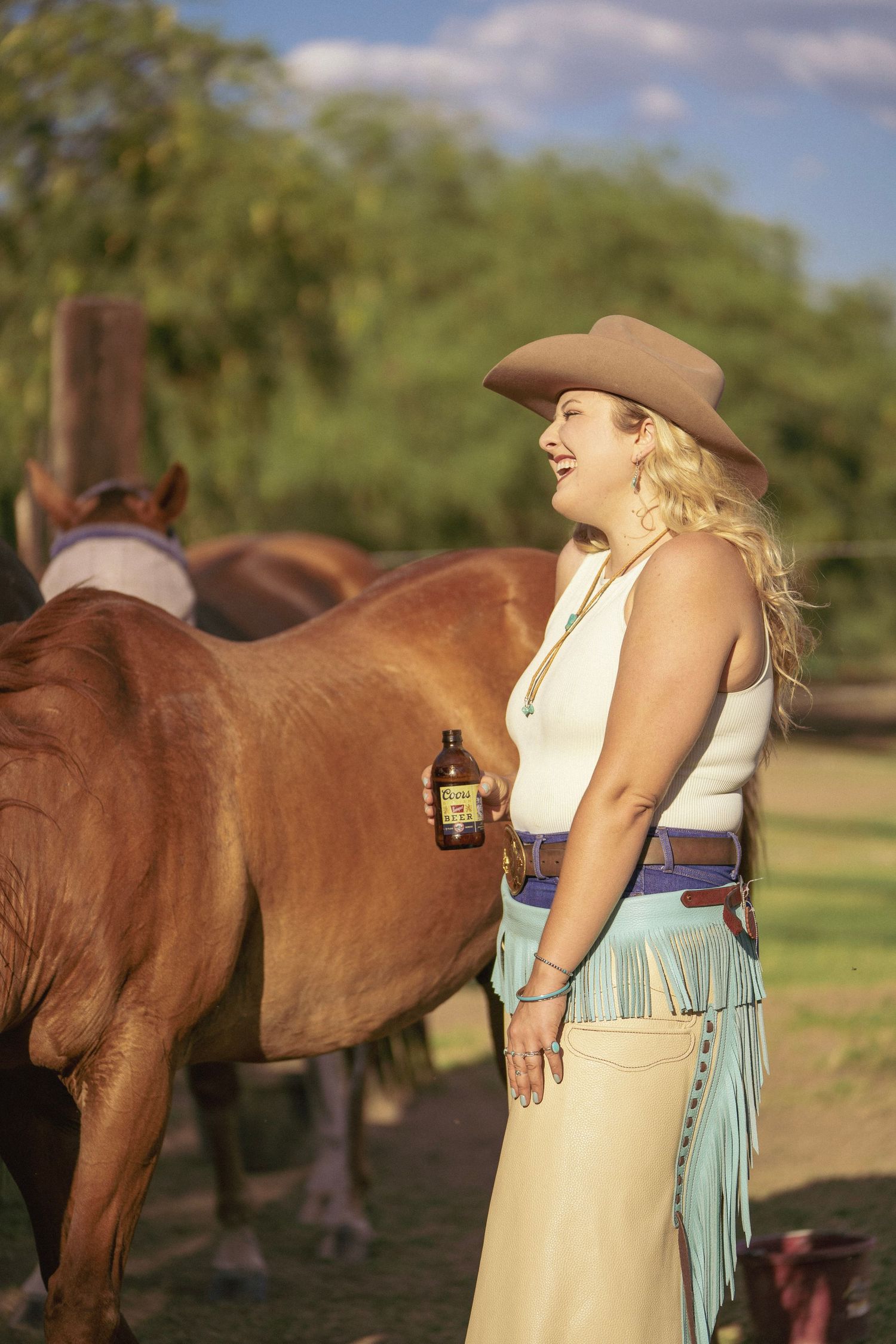 Cowgirl drinking a Coors Banquet beer while standing beside her horse in a western ranch setting.