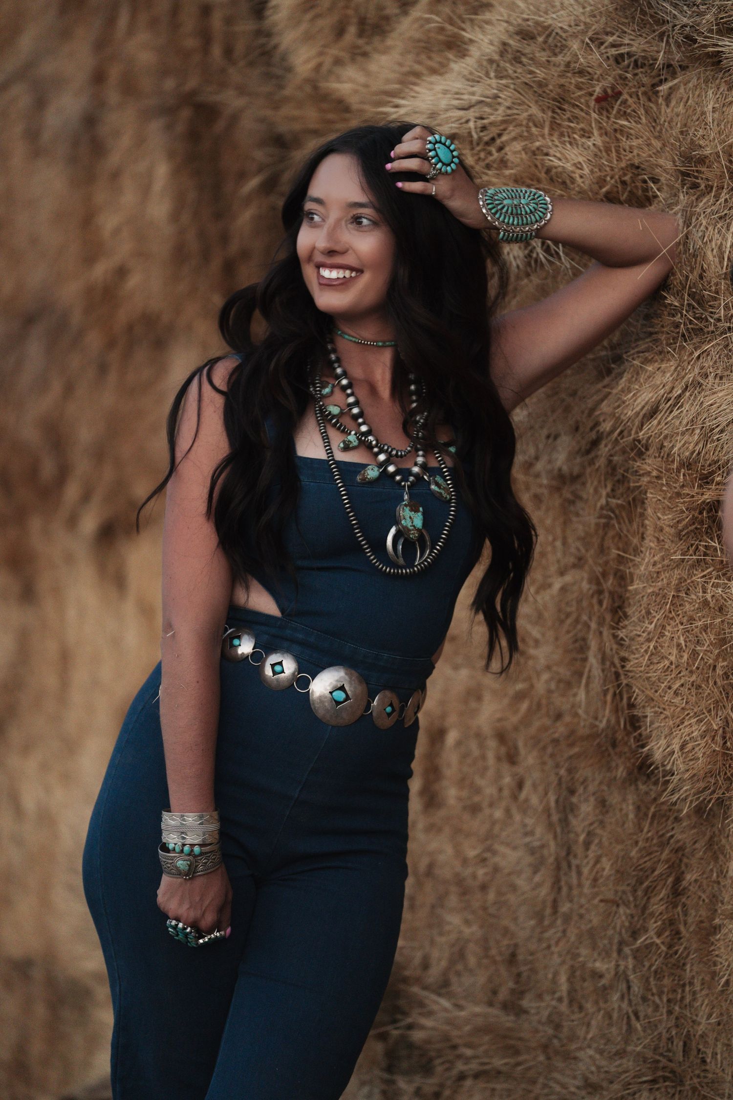 Girl leaning against a hay bale wearing a denim outfit and layered turquoise jewelry during a western lifestyle photoshoot.