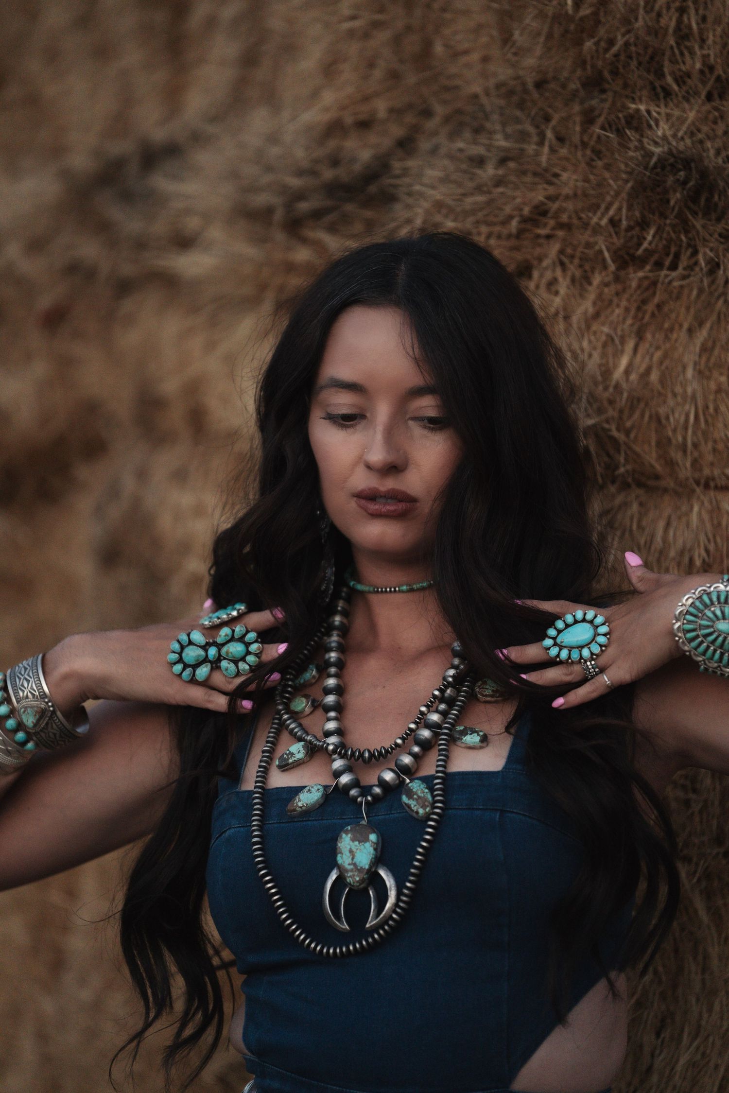 Girl leaning against a hay bale wearing a denim outfit and layered turquoise jewelry during a western lifestyle photoshoot.