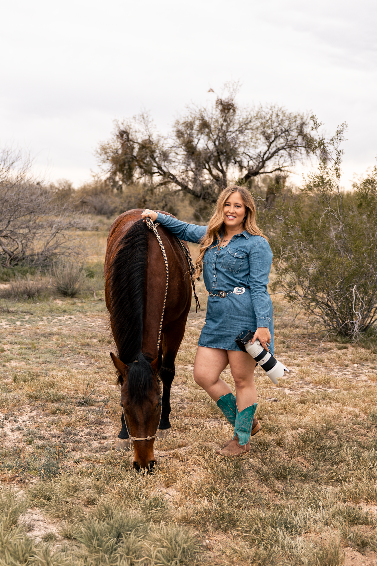 Western photographer in dress and holding camera beside bay horse in Arizona desert at sunset