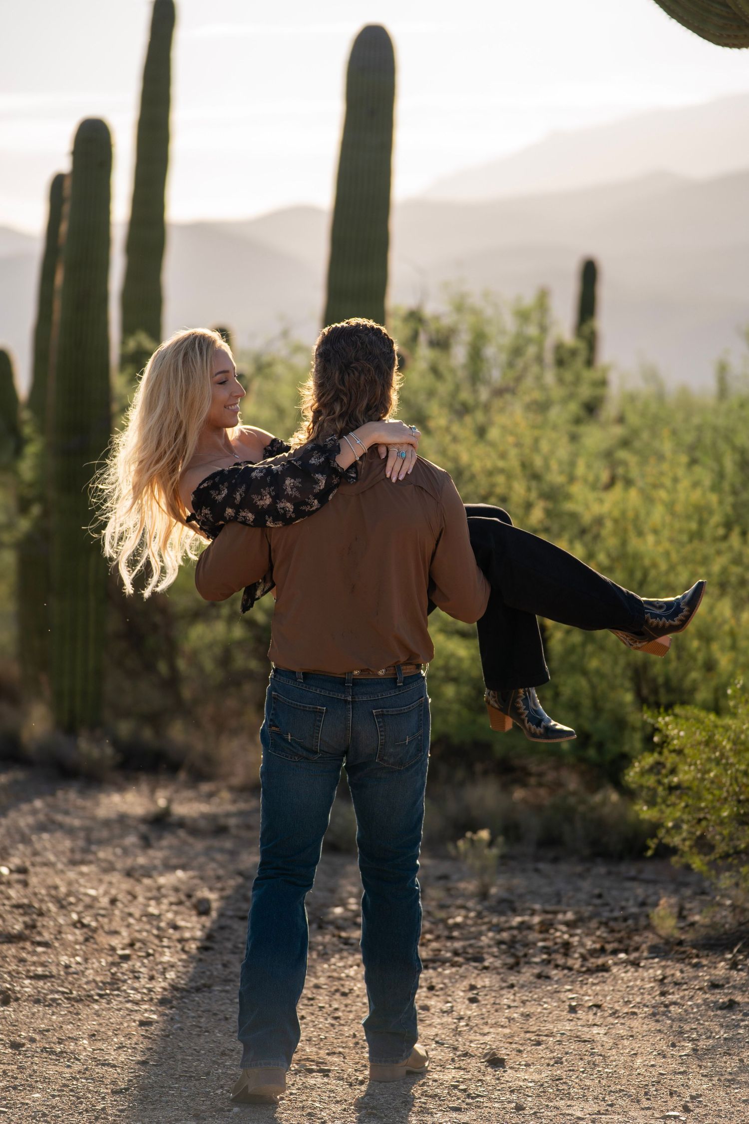 Man carrying his partner in his arms while walking away from the camera through the Arizona desert; the woman looks back toward the camera.