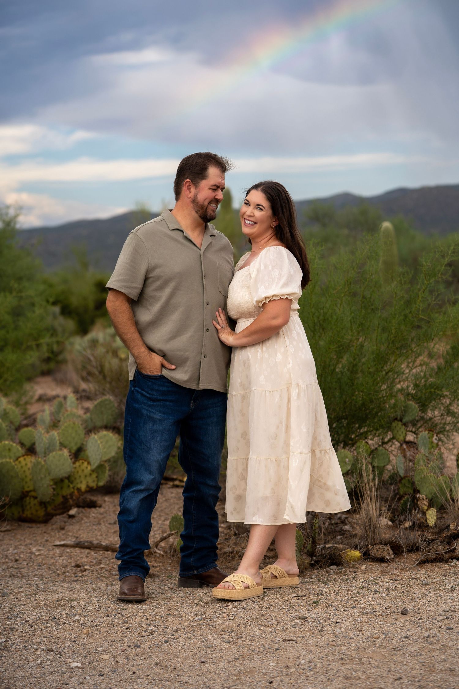 Couple standing together in the desert with a rainbow and dramatic clouds in the background.