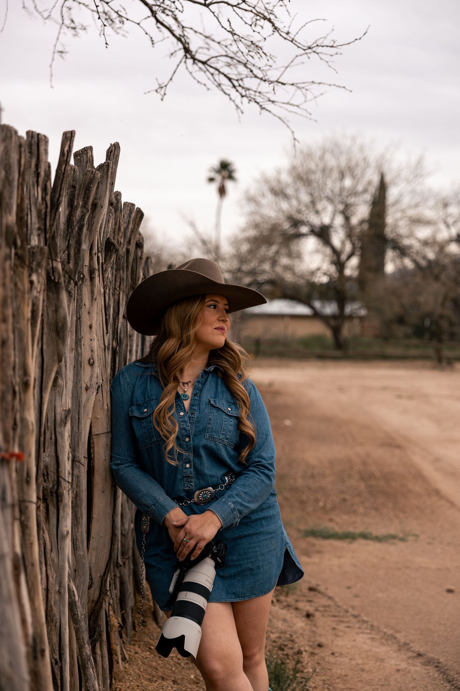 Western photographer in cowboy hat holding camera by rustic wooden fence in Arizona.