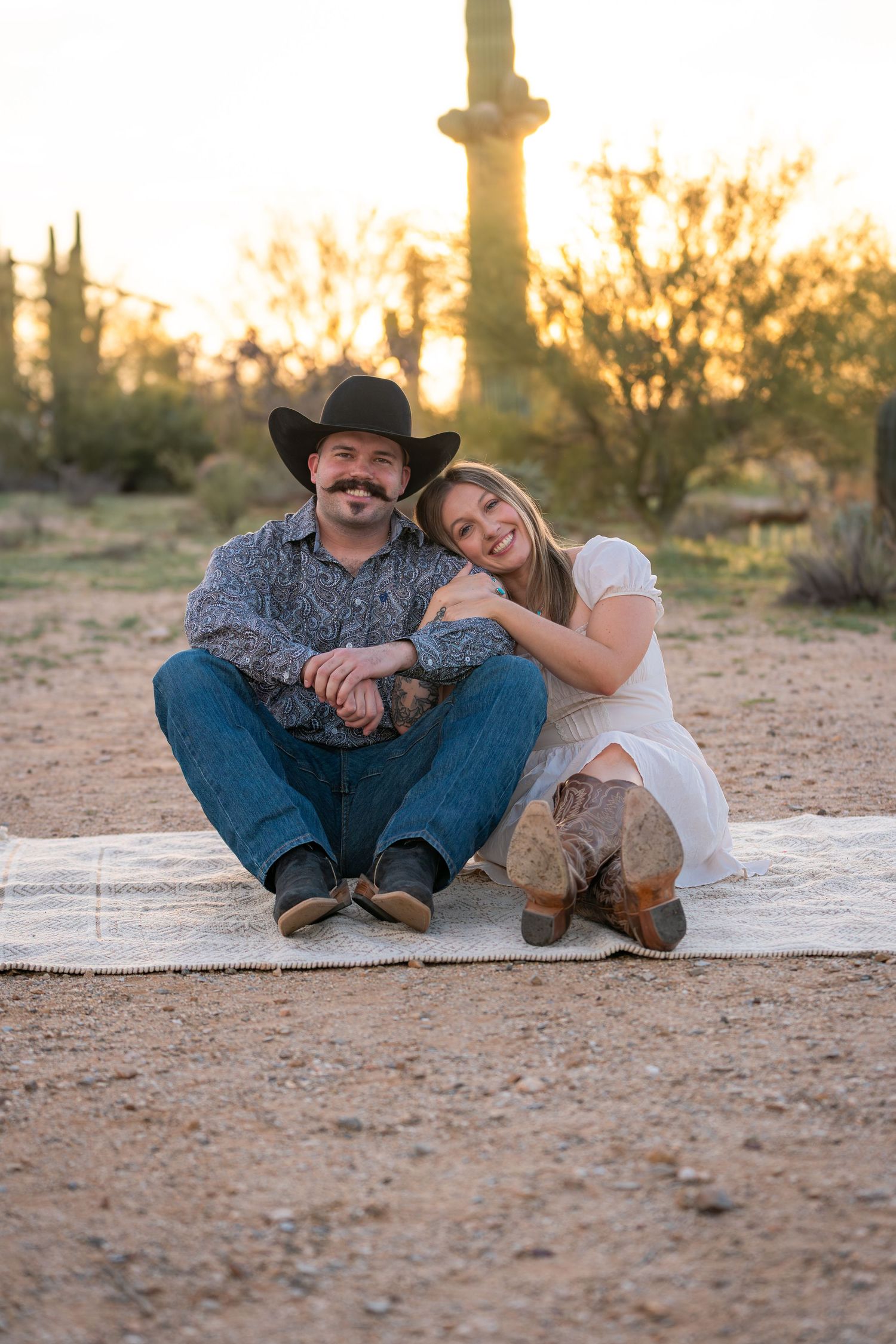 Western couple sitting on blanket at sunset in Arizona desert with saguaro cactus, romantic engagement photography