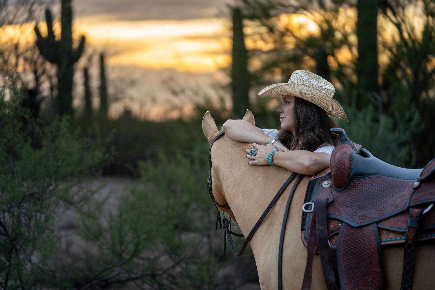 Cowgirl leaning against her horse in a warm desert landscape at sunset.