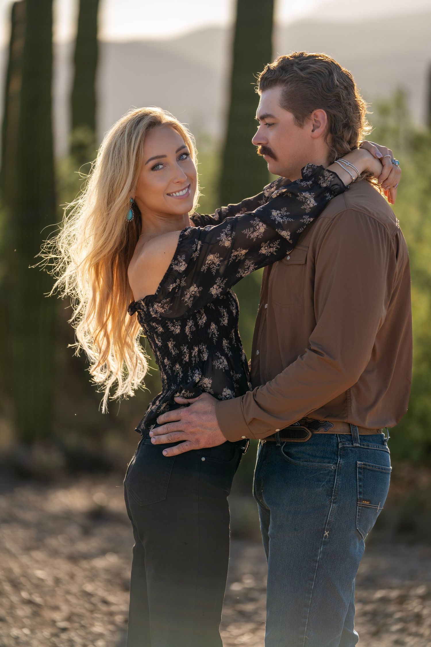 Woman with her arms around her partner’s neck, looking at the camera while he gazes at her during a western couple photoshoot in the desert.