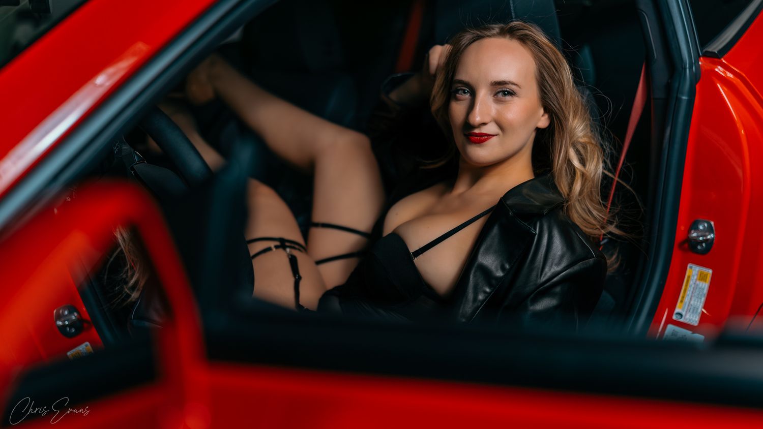 A person poses in a red car interior wearing a black top and stylish accessories.
